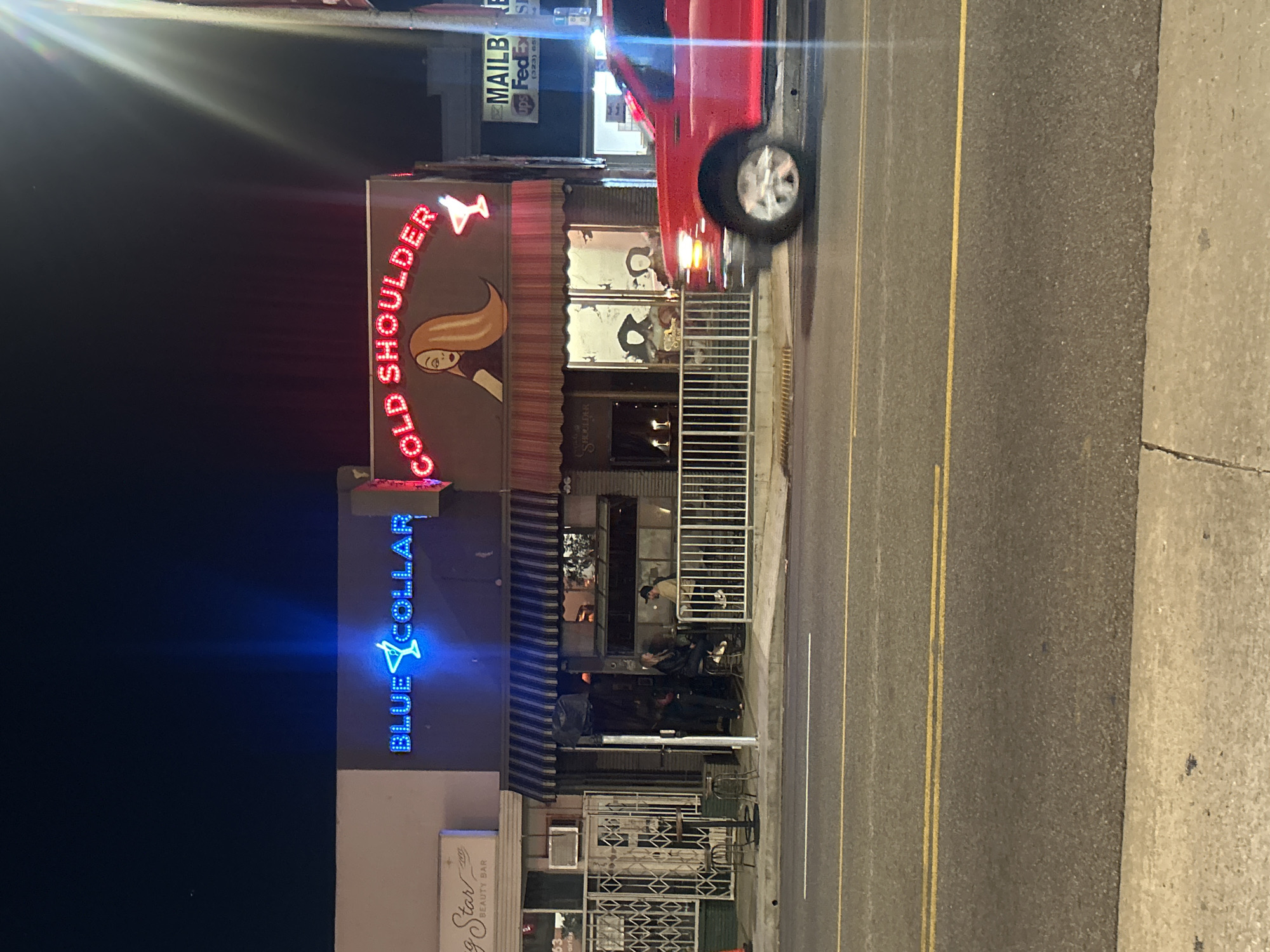 The image depicts a street scene at night, featuring storefronts with neon signs. One storefront is labeled "Blue Collar," with a blue neon sign above. Another storefront is labeled "Cold Shoulder," with a red neon sign depicting a woman and a cocktail glass. Several people are standing outside the "Cold Shoulder" establishment. A red vehicle is partially visible, positioned on the street adjacent to the storefronts. A street with double yellow lines and a sidewalk are also visible. Based on the OCR, it seems the location could be in the city of Los Angeles.