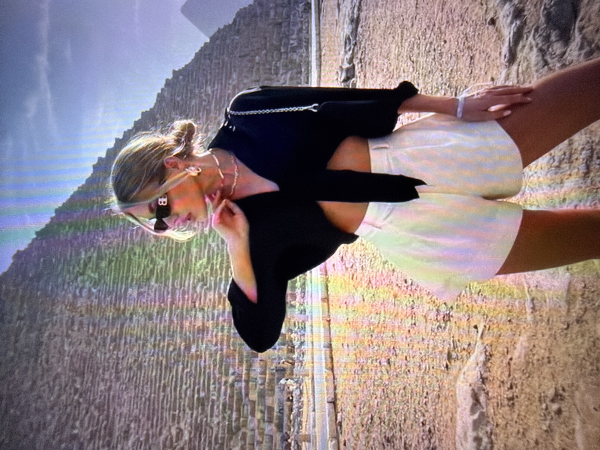 The image shows a woman in the foreground standing in front of the pyramids. She is wearing sunglasses, a black tied shirt, and white shorts. The pyramids are made of stone blocks and are under a blue sky. The ground is sandy. The image is a photograph that appears to have been taken on a digital camera, with some artifacts present. The image does not depict any scene in the city of Beșalma, Moldova.