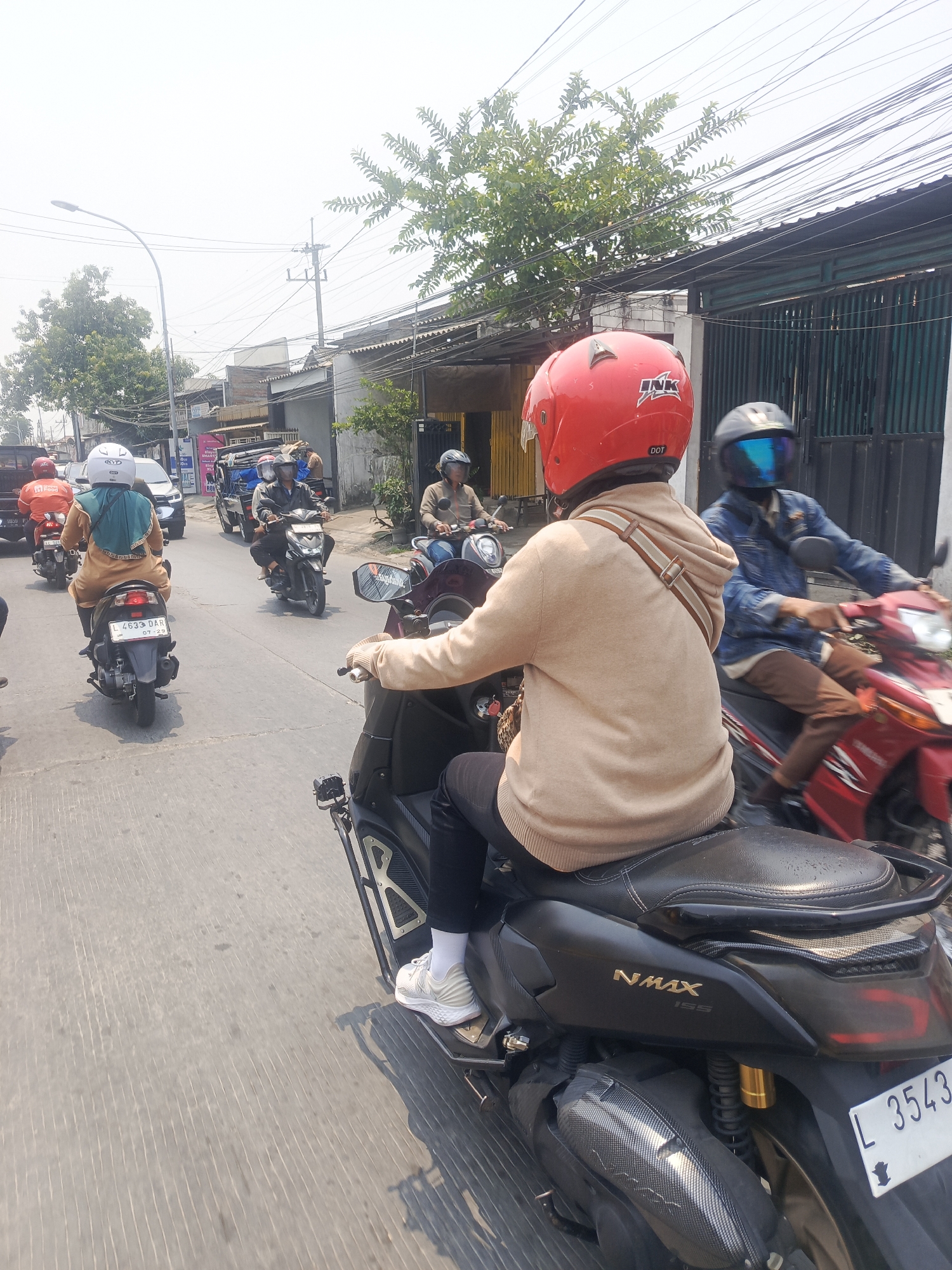 The image depicts a street scene in Surabaya, Indonesia, characterized by a bright, hazy sky and a road with visible white lane markings. The foreground is dominated by the rear view of a person riding a black NMAX 155 scooter, wearing a tan hooded sweatshirt and white sneakers. The rider's red helmet is prominently displayed. The scooter has a license plate reading "L 3543."
Several other motorcycles and riders are visible in the midground, moving in the same direction. One rider on a red scooter wears a blue denim jacket and brown pants. Further back, a rider in a tan shirt is on a motorcycle, and another person in a green scarf and tan outfit is on a scooter with a white helmet. The surrounding environment includes buildings with corrugated metal roofs and fences, as well as trees and power lines, typical of an urban setting. The overall impression is a busy, sunny day on a street in Surabaya.