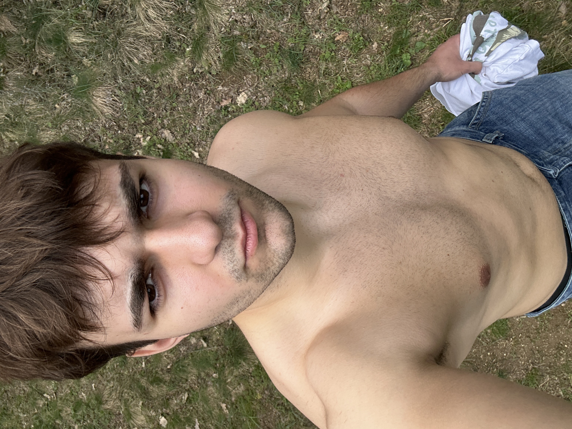 A shirtless man wearing blue jeans is positioned outdoors. He is holding a white t-shirt with a camouflage design. The background appears to be grass and vegetation. There is no indication of the city of Luliak, Bulgaria in the image.