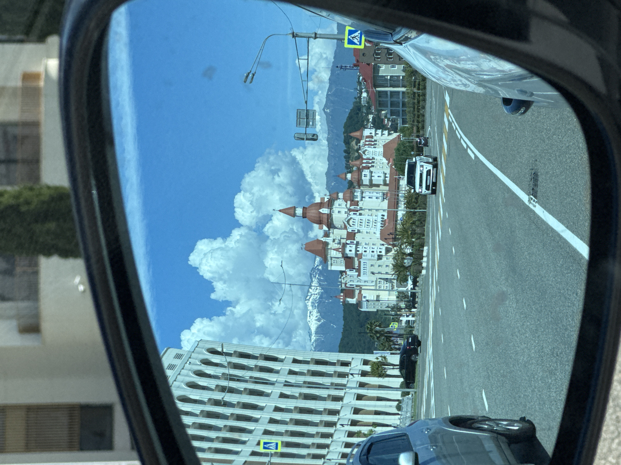 The image captures a reflection in a car's side mirror, showcasing a vibrant street scene and distant landscape. The reflection depicts a road running from the foreground towards the background, marked with white dashed and solid lines. Parts of the vehicle from which the photo is taken are visible on the right side of the mirror, including its bodywork and a wheel.
In the mid-ground of the reflection, a prominent white van with a distinctive front grille is seen on the road, followed by a dark sedan further away. Along the left side of the reflected road, an array of large, multi-storied buildings with white facades, numerous red-tiled peaked roofs, and small towers create an ornate, castle-like architectural style. Palm trees are scattered among these buildings. Beyond this developed area, majestic snow-capped mountains rise under a bright blue sky, adorned with large, fluffy white cumulus clouds. A street light pole with multiple lamps and a blue pedestrian crossing sign are also visible in the reflection. In the lower-left section of the mirror, another large building with a series of arched windows or balconies can be seen.
The scene unfolds under clear, sunny daytime conditions, indicative of pleasant weather. The unique combination of resort-style architecture, palm trees, and the backdrop of snow-capped mountains strongly points to a location like Sirius, Russia, which is situated on the Black Sea coast near the Caucasus Mountains. The image captures a momentary view from a journey, presenting a picturesque environment through an unconventional perspective. No discernible alphanumeric text is visible, only the universal symbol for a pedestrian crossing.