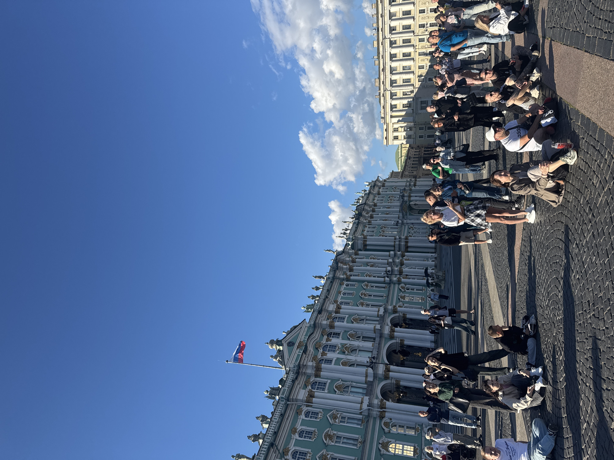 A group of people are sitting on the cobblestone ground in front of a grand, ornate building. The building is light green with white columns and gold accents, featuring many windows and statues along its roofline. A Russian flag flies from a flagpole on the building's facade. The sky is a clear, vibrant blue with scattered white clouds. The sun is bright, casting distinct shadows, suggesting it is daytime, likely late morning or afternoon. The people are dressed in casual, modern clothing. Some are looking towards the building, while others are interacting with each other or looking at their phones. The overall atmosphere appears relaxed and leisurely. The scene is set in a public square or plaza, likely a tourist attraction given the architecture and the presence of a flag. There is no discernible text visible in the image.