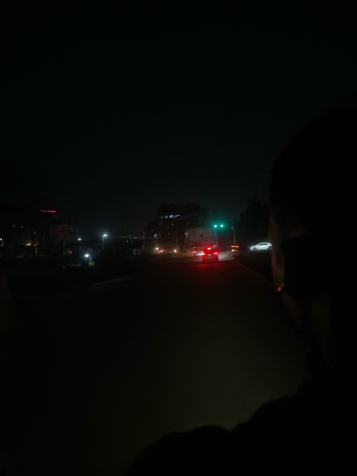 The image captures a nighttime scene on a street in Abuja, Nigeria. The foreground is dark, with the silhouette of a person's head and shoulder partially visible on the right side, suggesting the photo was taken from a moving vehicle or from behind someone. The street ahead is illuminated by streetlights and the headlights and taillights of cars. Several vehicles are visible on the road, including a white car in the distance, and red taillights of other vehicles. A traffic light with a green signal is illuminated on the left side of the road. Buildings with some lights on are visible in the background, hinting at an urban environment. The overall atmosphere is dark and somewhat blurry, typical of a nighttime, low-light photograph.