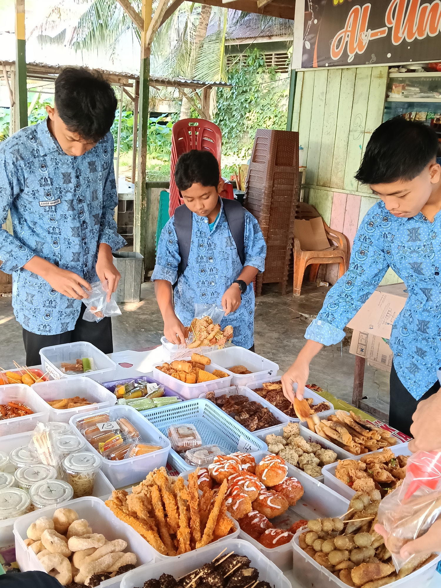 Three young men wearing matching blue patterned shirts are standing around a table laden with various foods. The food is displayed in clear plastic containers and includes items such as fried snacks, skewers, and pastries. The young men are interacting with the food, placing it into plastic bags. The background includes a wooden structure, stacked chairs, and greenery, along with a sign in script behind the table. The location appears to be an outdoor food stall, possibly in Lhokseumawe, Indonesia.