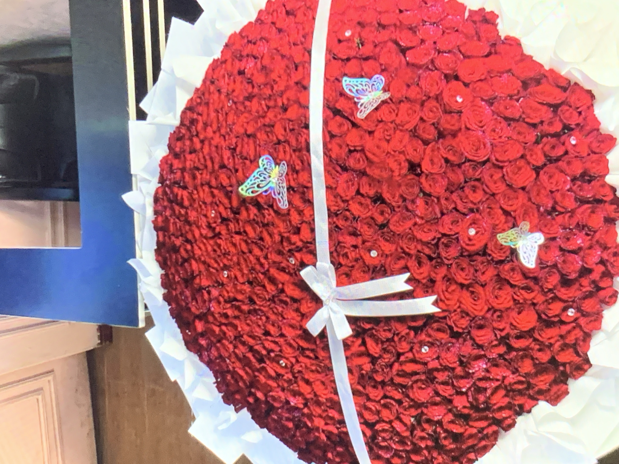The image shows a large, heart-shaped bouquet composed entirely of red roses. The roses are tightly packed together, and the bouquet is decorated with three butterfly embellishments and small, scattered rhinestones. A white ribbon is wrapped around the bouquet, tied in a bow near the center. The bouquet is surrounded by white tissue paper. The bouquet is partially in front of a cupboard or wall. There is no evidence in the photo to prove location in the city of Rafin Par, Nigeria.