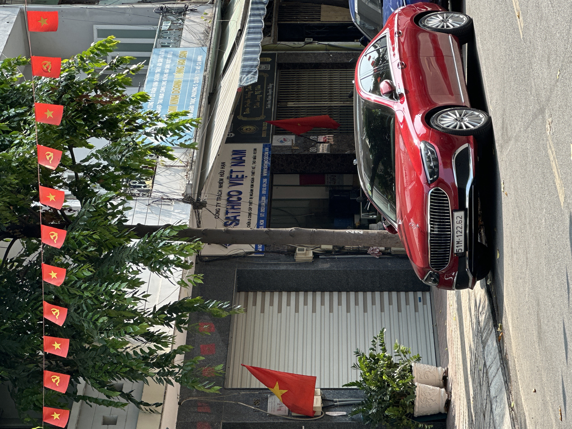 The image shows a street scene in Ho Chi Minh City, Vietnam. A red car is parked along the side of the road. A tree with a string of Vietnamese flags is visible on the left. Several buildings are visible, including one with a sign for "SATHICO VIET NAM". The license plate number on the car is 51M-122.62. There are flowerpots with small plants near the building.