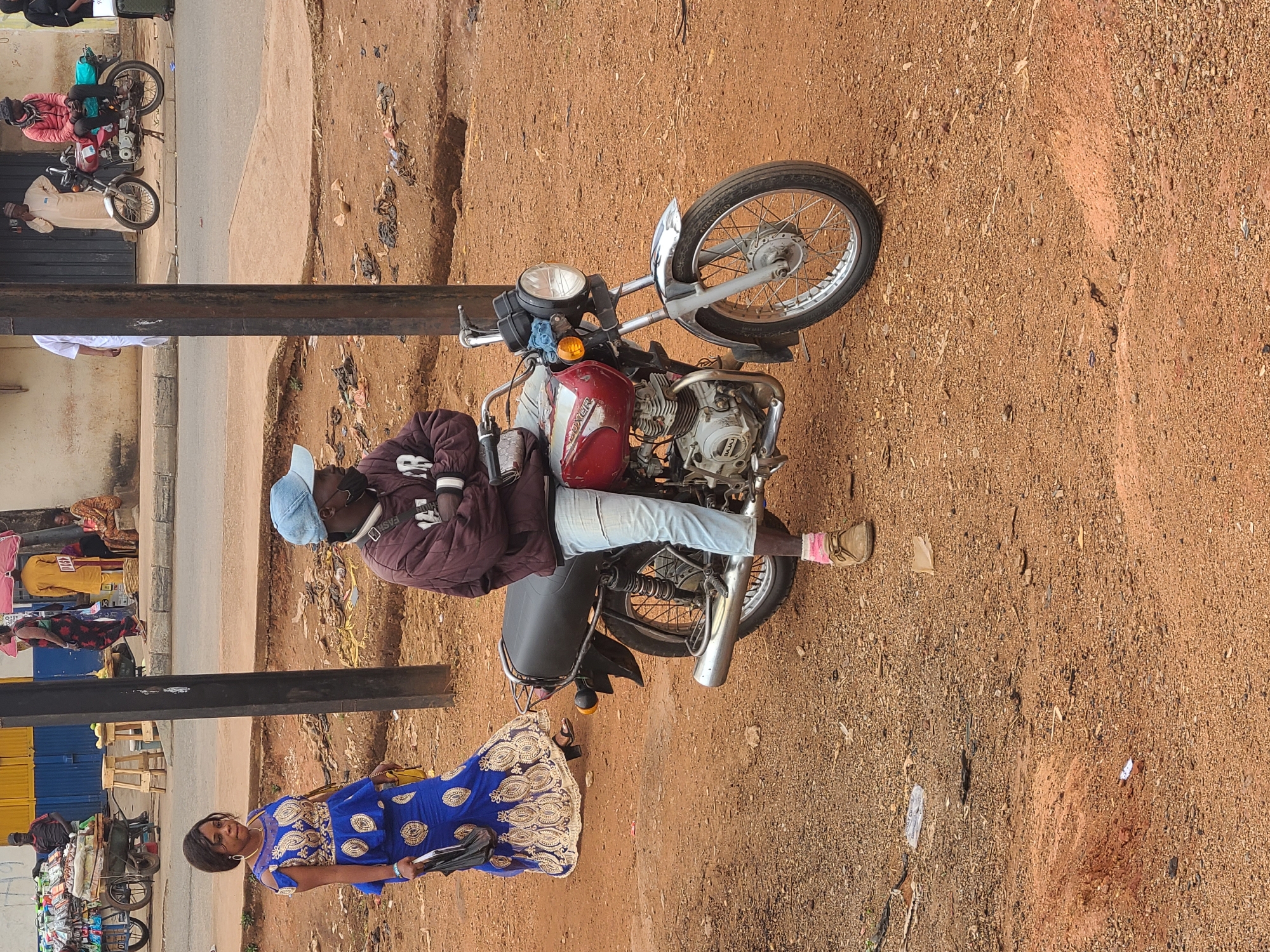 The image shows a street scene in Jos, Nigeria. A man in a burgundy jacket, blue jeans, and a blue baseball cap is sitting on a red and silver motorcycle parked on a dirt patch alongside a paved road. A woman in a blue patterned dress is walking past the motorcycle. In the background, there are market stalls with various goods and people. Debris and trash are scattered on the ground near the motorcycle. A metal pole is visible in the foreground.