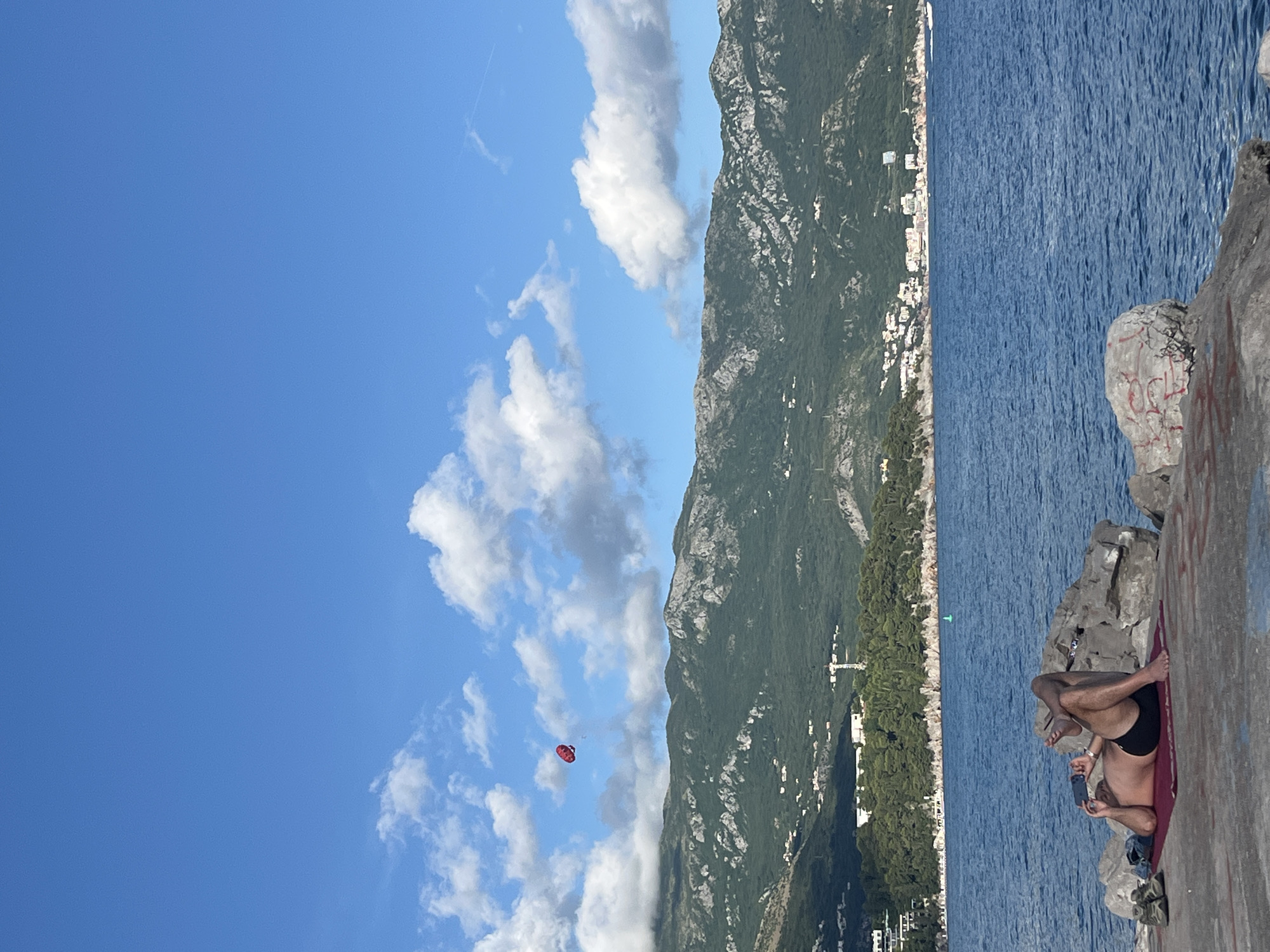 This vibrant image captures a sunny coastal scene, likely Budva, Montenegro, during daylight hours. A clear blue sky, dotted with fluffy white and grey clouds and a faint airplane contrail, dominates the upper left portion of the frame.
A prominent green mountain range, thickly covered in vegetation, stretches across the midground, its peaks reaching into the sky. Along the lower slopes of these mountains, numerous white buildings of a town are visible, nestled along the coastline.
The deep blue sea fills the right and lower-middle sections of the image, displaying a textured surface from ripples. In the distance, above the water, a bright red parasail with a person underneath is visible against the sky, indicating a recreational activity.
In the foreground, on a concrete or rocky platform by the water's edge, a person is relaxing. They are lying on their back on a reddish mat or towel, wearing dark swim shorts, with their legs bent. They are holding a smartphone in their hands and appear to be looking at the screen. The concrete surface directly beside them is marked with red graffiti, with some letters visible. The scene suggests a leisurely day by the sea, with clear weather.