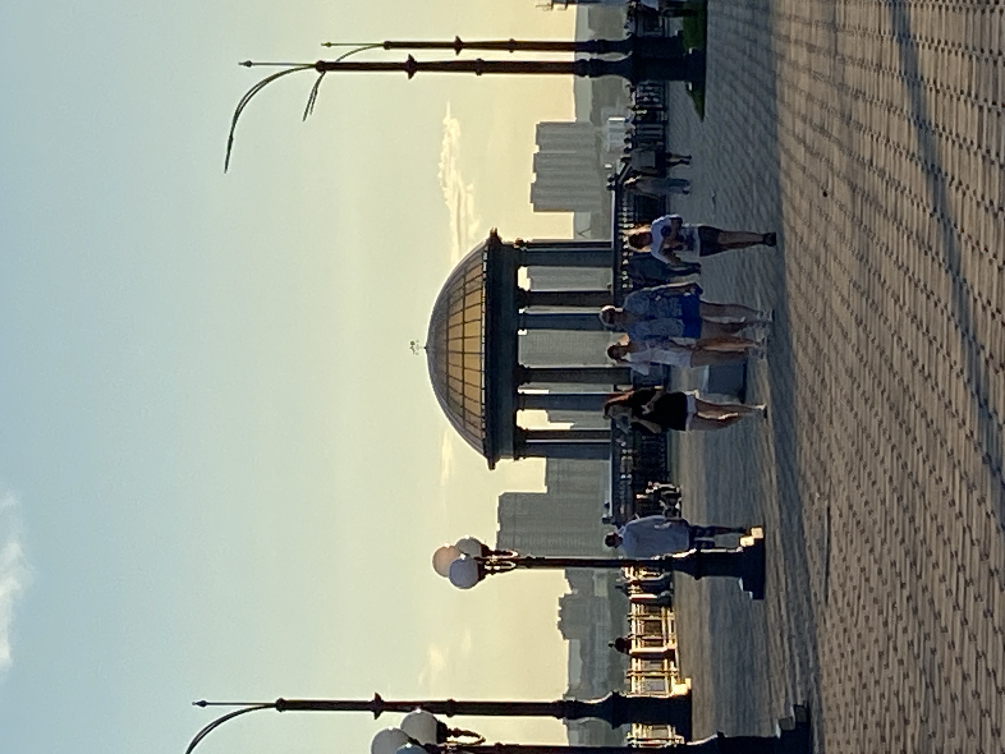 A scenic view of the Blagoveshchensk embankment along the Amur River at dusk. The sky is a soft gradient of blue and yellow, with a few wispy clouds. In the foreground, a paved walkway is lined with lampposts, some with multiple globe-shaped lights. Several people are strolling along the embankment. A prominent feature is a classical-style rotunda with a domed roof, surrounded by columns. In the background, the cityscape of Blagoveshchensk, Russia, is visible with its modern high-rise buildings. The overall atmosphere is peaceful and recreational.