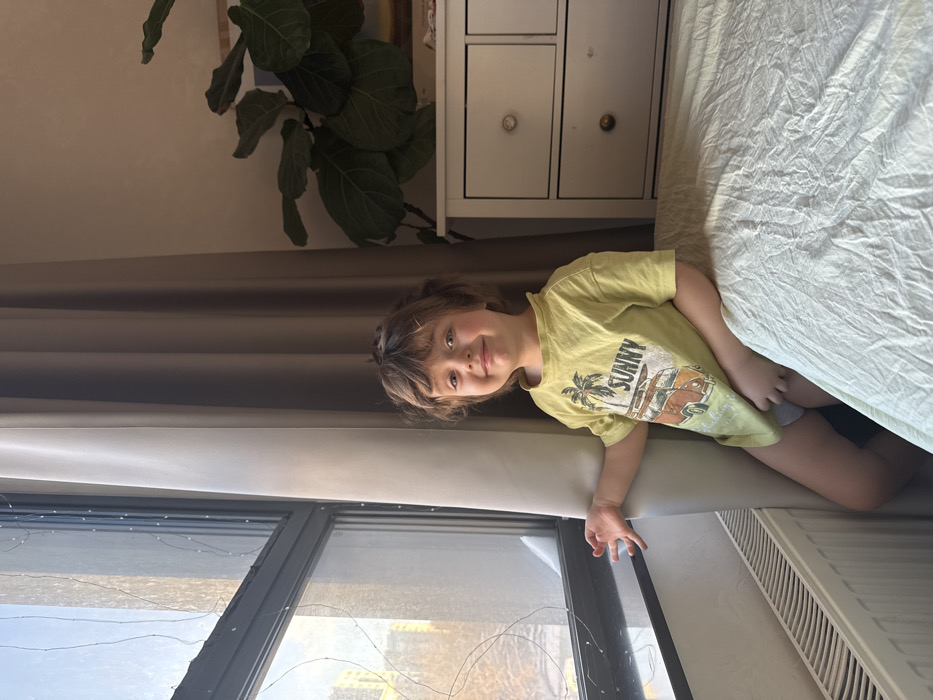 A young boy with brown hair and a yellow "Sunny" t-shirt smiles towards the camera. He is positioned next to a window with drawn curtains, and a heater is visible beneath his legs. Above the boy, a white nightstand with drawers is seen along with a green leafy plant. The upper right corner reveals a white bed covering.
