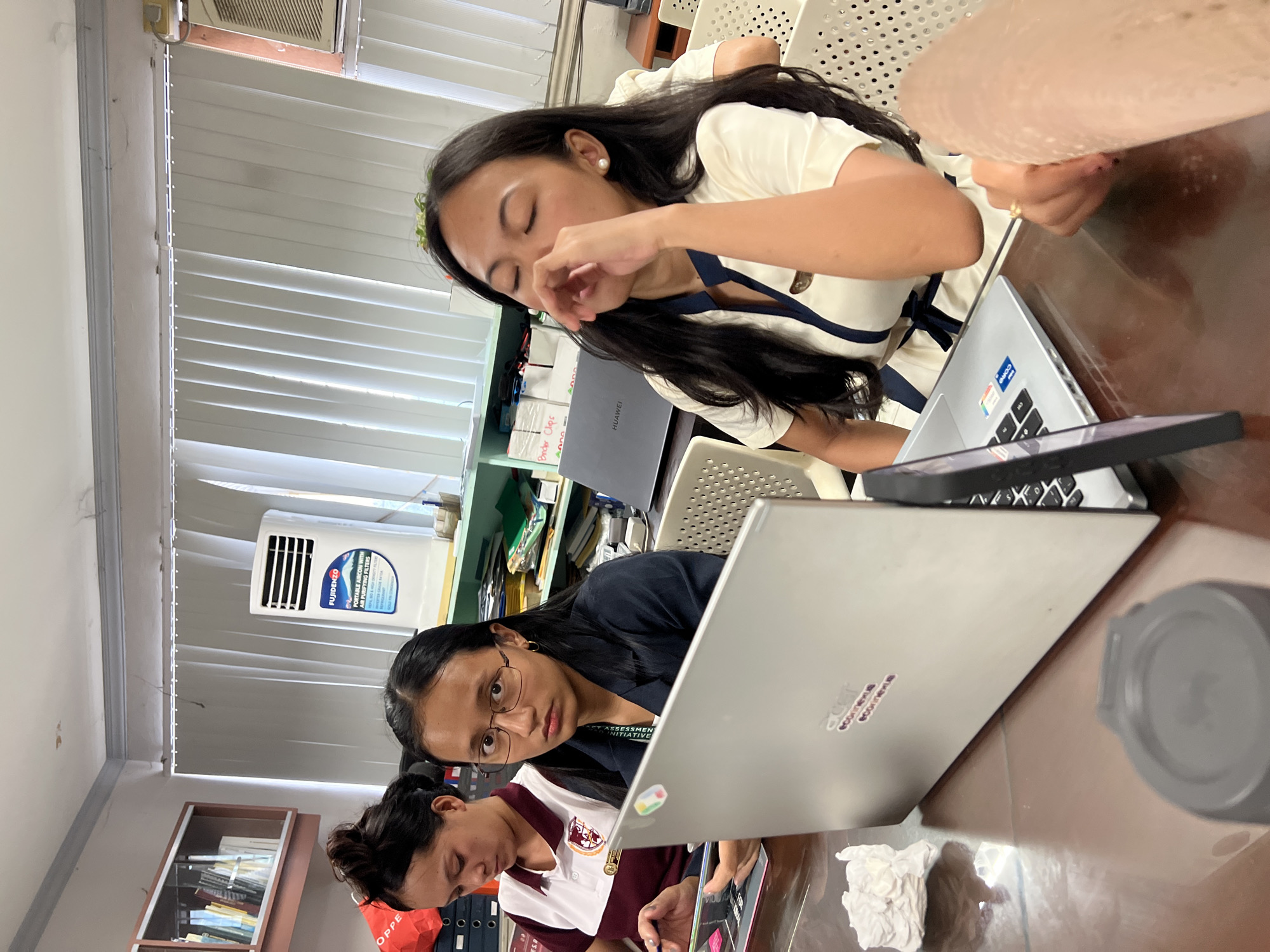 The image depicts a scene indoors, likely an office or study area, with three young women engrossed in their work. The setting appears to be a classroom or a collaborative workspace, as indicated by the presence of multiple laptops and a desk. The lighting suggests it is daytime.
The woman in the foreground, wearing glasses and a dark top, is actively engaged with a silver laptop. Her gaze is directed towards the screen, suggesting focus and concentration. To her left, another woman in a white and navy outfit is also looking down at a laptop, her eyes closed and hand near her face, possibly indicating fatigue or deep thought. A third woman, partially visible in the bottom left, is hunched over a laptop as well, her attention seemingly captured by the device.
On the desk, several objects are present, including laptops, a smartphone, crumpled paper, and a reusable cup. A visible piece of text on a laptop says "HUAWEI". Another laptop has stickers that read "econnexue". There is also a Fujidenzo portable aircon visible in the background with text mentioning "PORTABLE AIRCON WITH AIR PURIFYING FILTERS". Behind the subjects, shelves are filled with folders and binders, adding to the impression of an academic or professional environment. The women appear to be students or employees working on tasks that require their full attention. The overall mood is one of quiet industry.