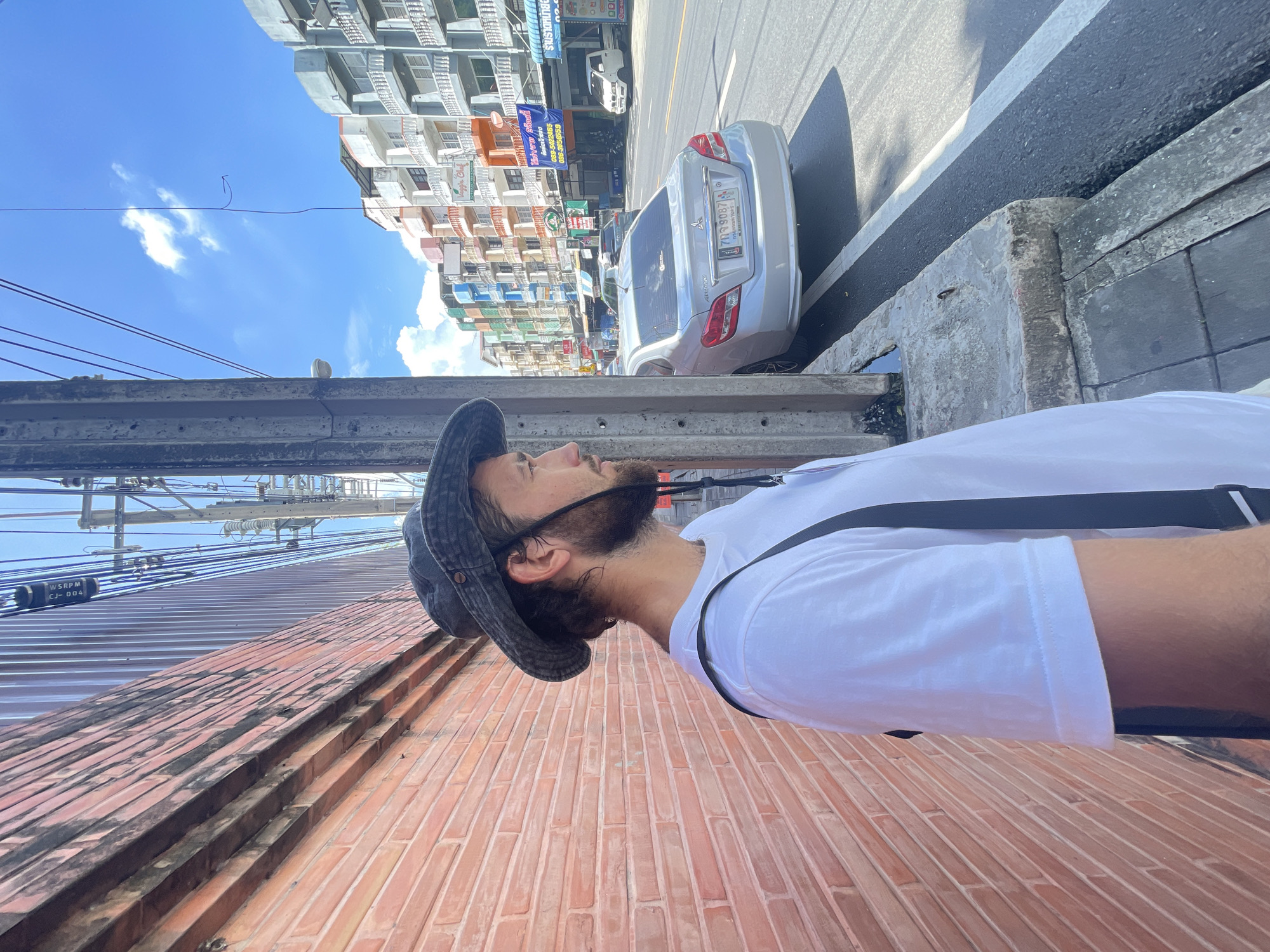 A man stands on a sidewalk in Bangkok, Thailand, looking to his right. He wears a dark bucket hat and a white t-shirt, with a black strap crossing his chest. The sidewalk is made of concrete, with a raised edge. Behind him is a brick wall. To his right, a street runs parallel to the sidewalk, with a silver car driving on it. Buildings line the street, with colorful facades and balconies. Utility poles and wires are visible against a bright blue sky with scattered clouds. The scene appears to be during the daytime, under sunny conditions.