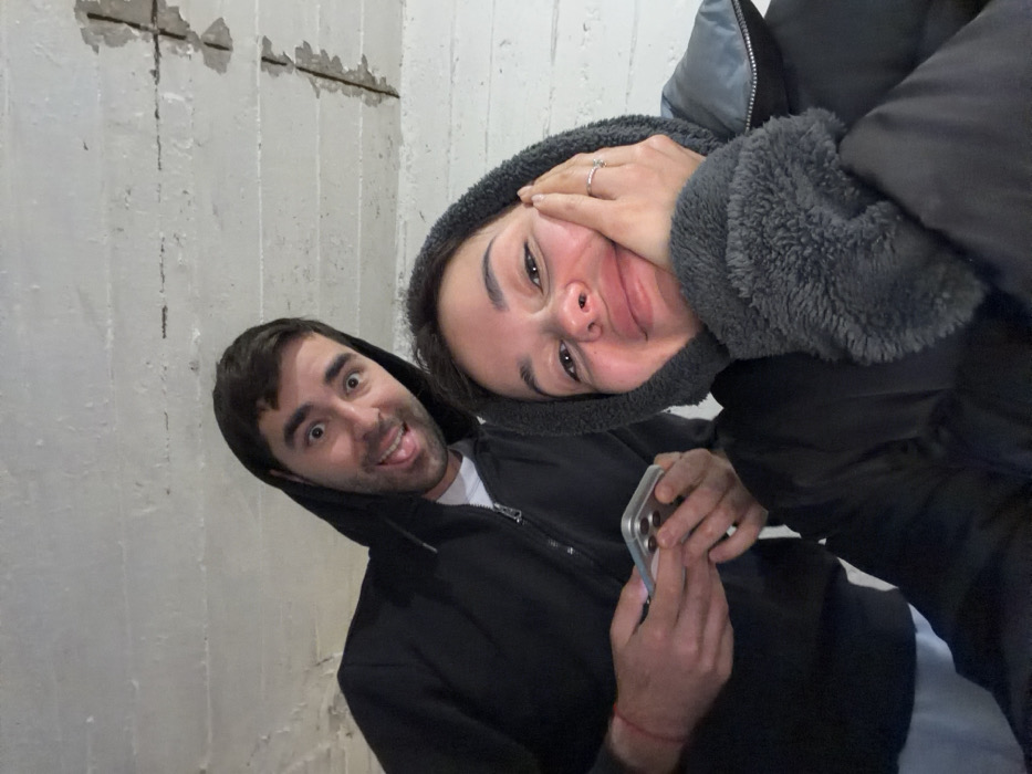The image depicts a close-up, selfie-style photograph of two individuals, an adult female and an adult male. The female, positioned in the foreground on the right, has fair skin and dark hair, and is wearing a dark puffer jacket with a grey fleece-lined hood and cuffs. She is smiling broadly, with her left hand, adorned with a silver-toned ring, resting against her cheek. The male, positioned in the background on the left, has fair skin and dark hair, and is wearing a dark hooded sweatshirt over a white t-shirt, with a red string bracelet on his right wrist. He is looking towards the camera with a wide-eyed, open-mouthed expression, his tongue slightly extended. He is holding a light-colored smartphone, featuring a visible triple camera array on its rear, in both hands. The background consists of an unfinished, light-colored concrete ceiling or wall. This surface exhibits visible horizontal lines, textured concrete, and several areas where the concrete has chipped away, exposing corroded steel reinforcement bars. The scene is located in the city of Tel-Aviv, Israel.