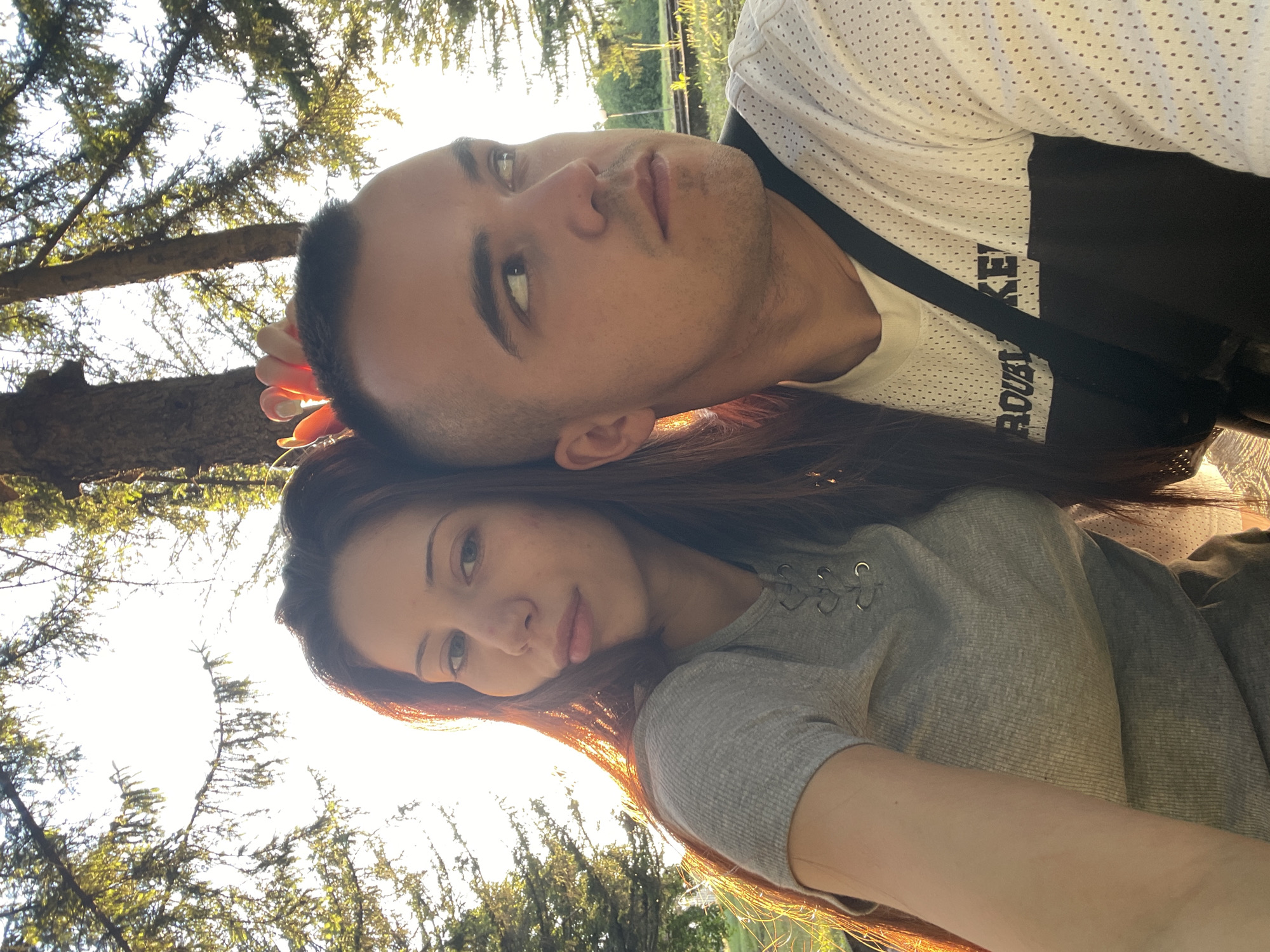 A couple is captured in a close-up selfie outdoors, surrounded by trees and bathed in warm sunlight. The woman on the left, with long, dark, reddish-brown hair, gazes directly at the camera with a neutral expression. She is wearing a grey, ribbed t-shirt with a lace-up detail at the neckline. To her right, a man with a closely cropped haircut looks away from the camera, his profile visible. He is wearing a white t-shirt with black accents and a black strap across his chest. The background shows dappled sunlight filtering through green foliage, suggesting a park or wooded area. The overall impression is a candid moment of connection between the two individuals in a natural setting.