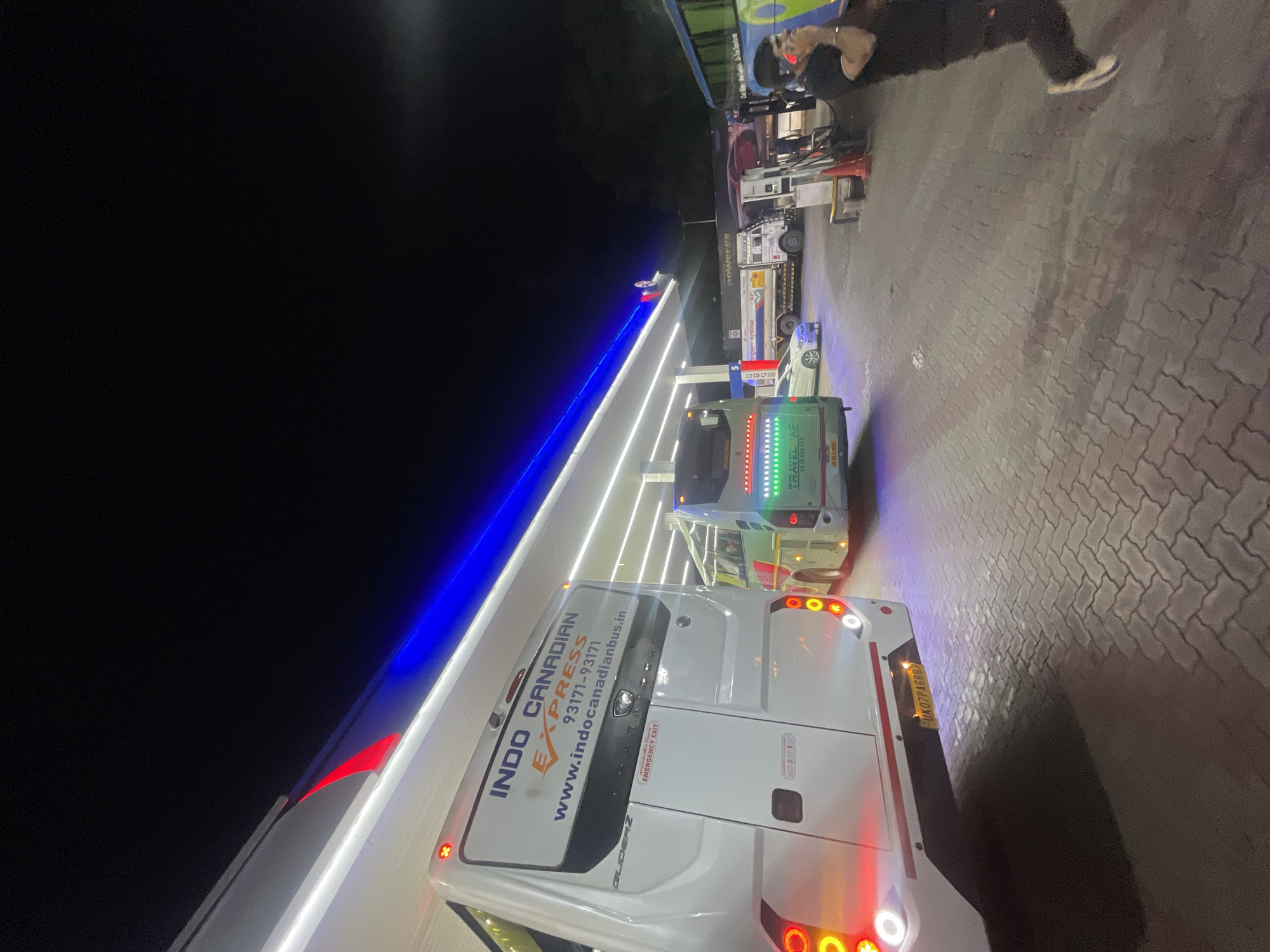 The image depicts a nighttime scene at a gas station in Kajheri, India. The foreground features the rear of a white bus with "INDO CANADIAN EXPRESS" branding and contact information. To the right of this bus, another bus is visible, also with illuminated signage. In the middle ground, a white car is parked, and a truck with "HP Power95" branding is also present. The gas station features blue and white illuminated structures overhead. In the background, several other vehicles and what appear to be fuel pumps are situated. A person dressed in dark clothing is on the right side of the image, talking on a mobile phone. The ground is paved with interlocking bricks, and shadows are cast across the scene.