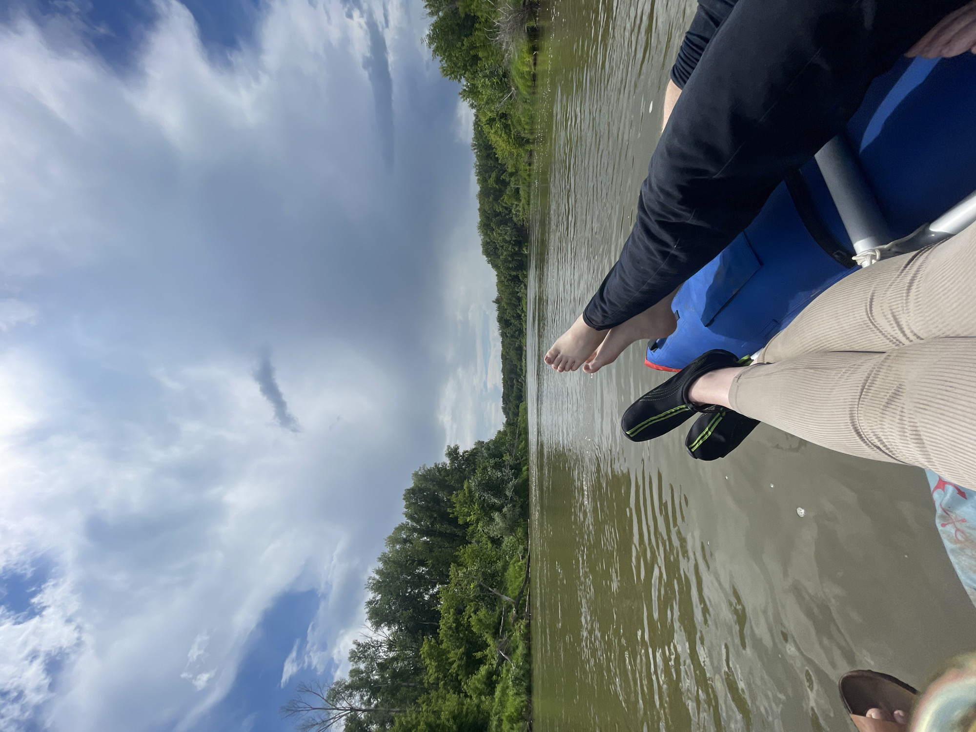 The image captures a serene scene on a river, likely the Irtysh River near the village of Samorodovo, Russia, given the characteristic landscape. Two individuals are relaxing in what appears to be an inflatable raft or kayak, their legs extended towards the camera. One person is wearing khaki-colored corduroy pants and a sandal on their left foot, while the other is clad in dark pants and black water shoes with green accents.
The raft, predominantly blue, rests on the calm, greenish-brown water of the river, which reflects the overcast sky and the lush, green foliage lining its banks. The riverbanks are densely wooded with deciduous trees, creating a natural, unspoiled vista. The sky is dramatic, with a mix of dark, heavy clouds and patches of brighter sky, hinting at changeable weather. Several small bubbles are visible on the water's surface, suggesting gentle movement or aeration. The overall atmosphere is peaceful and suggests a leisurely activity enjoyed in nature.