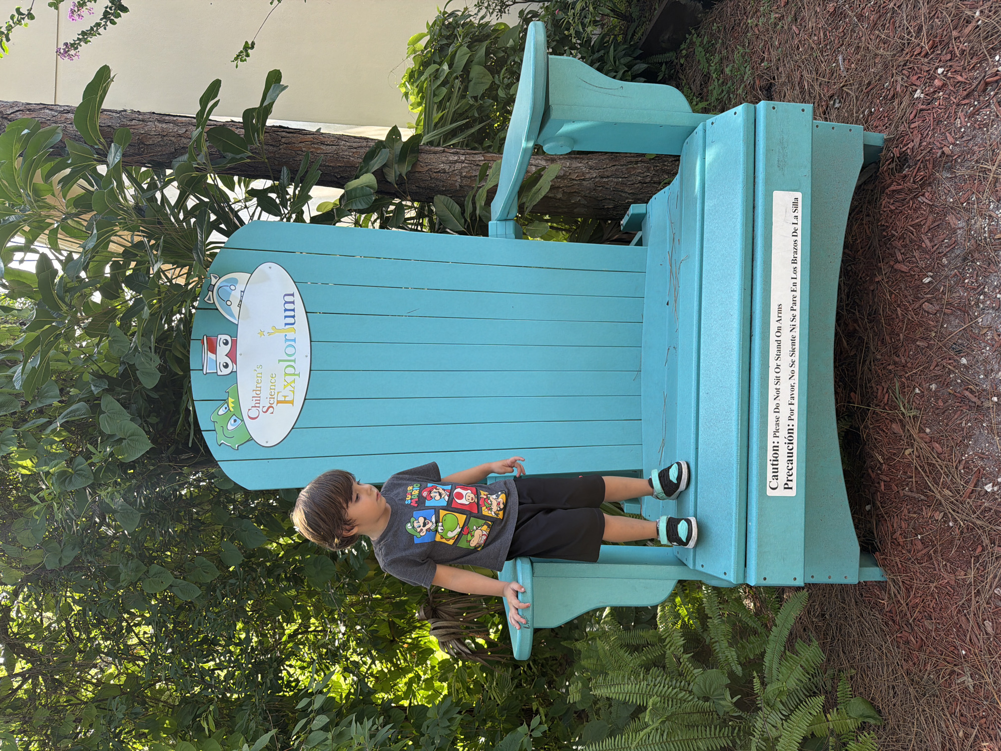 A young boy with brown hair stands on the edge of a large, turquoise Adirondack chair. He is wearing a gray t-shirt with images of Super Mario characters and black shorts. The chair is outdoors, situated in a grassy area with ferns and trees. A sign on the back of the chair reads "Children's Science Explorium". Another sign at the bottom of the chair states "Caution: Please Do Not Sit Or Stand On Arms" in both English and Spanish. The scene appears to be in Boca Raton, Florida, given the presence of the Children's Science Explorium, a science museum for children located in that city.