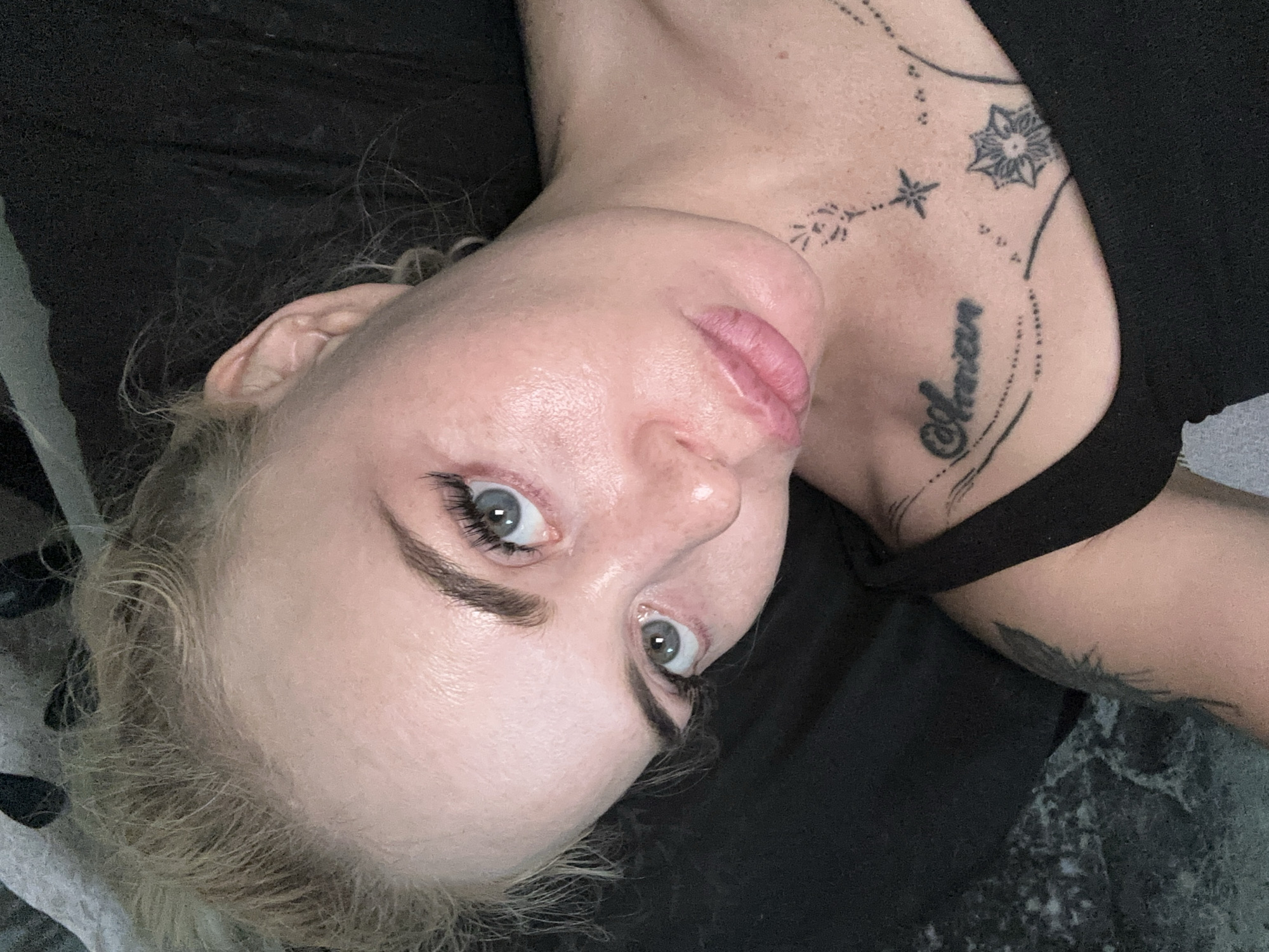 A medium close-up, high-angle, selfie-style shot shows a young, fair-skinned woman with blonde hair pulled back from her face, lying on her side against a dark, textured background. Her eyes are open and looking towards the camera, with bright blue irises. Her eyebrows are dark and well-defined, and her eyelashes appear to be enhanced. She has subtle pink lipstick on her lips, which are slightly parted. Her skin is visible with some texture and what appear to be light freckles or skin blemishes. She is wearing a black tank top, and the upper part of her chest and shoulder are visible. On her chest, there are several black tattoos. One tattoo on her collarbone reads "Annian" in cursive script, and other tattoos include abstract designs and what looks like a stylized flower or star. The lighting is soft, highlighting the contours of her face and chest. The background is predominantly dark and out of focus, suggesting an indoor setting. There is no indication of the city or location within the image itself.