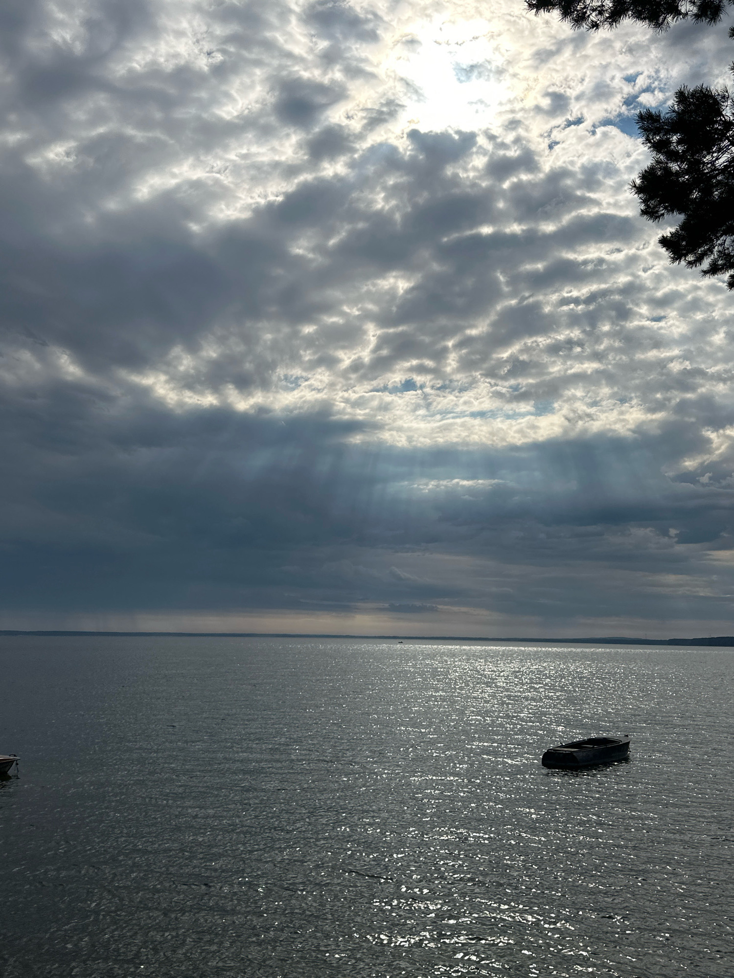 The image depicts a serene scene on a large body of water, likely Lake Naroch, located within the Naratski Sel'ski Sovet in Belarus. The sky is dramatically filled with a mix of heavy, dark clouds and patches of bright sunlight, creating visible sunbeams that pierce through the clouds and illuminate the water below. The water's surface is rippled, reflecting the sunlight in shimmering patterns. In the mid-ground, a solitary, dark-colored rowboat floats peacefully on the water. The horizon line is visible in the distance, with a faint outline of land under the cloud-laden sky. In the upper right corner, the dark silhouette of pine tree branches is visible, framing the sky and water. No people are visible in the frame.