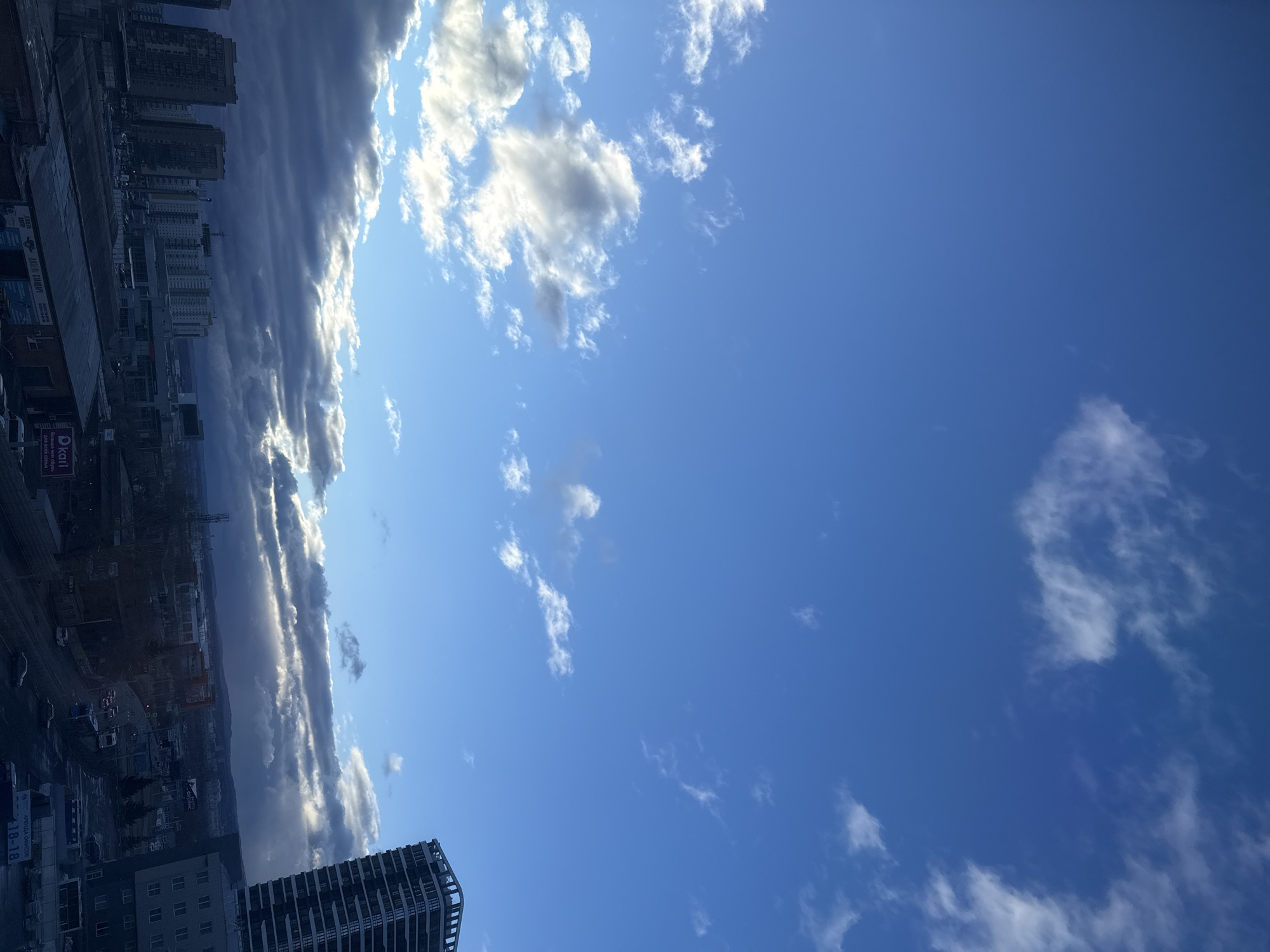 The image presents a cityscape under a blue sky with scattered clouds. Buildings of varying heights are visible, appearing denser in the left portion of the image. Some structures appear to be residential apartment buildings, while one prominent building is a high-rise with a modern architectural design. The lower left corner contains an area with some commercial signage. The sky occupies the majority of the frame and is a vivid blue with scattered white clouds, some appearing more prominent than others. Based on the urban characteristics visible, the image is in Krasnoyarsk, Russia.