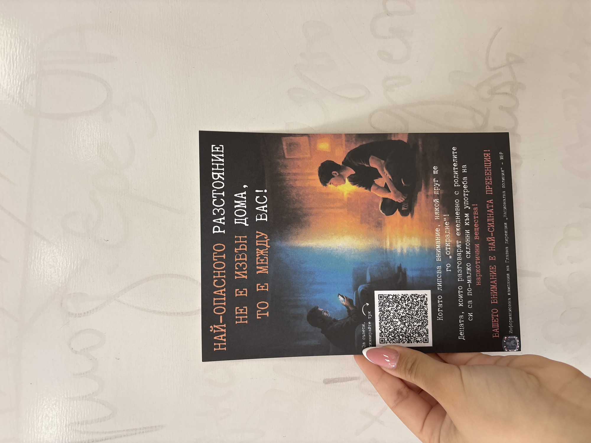 A female hand with light pink and white-tipped nails holds a vertical black flyer against a white surface with faint, watermark-like cursive patterns. The flyer, part of an information campaign by the General Directorate "National Police" of the Ministry of Interior in Bulgaria, addresses drug prevention and parental attention.
The flyer features a stylized image of two young men. One figure, illuminated by warm orange light, sits with his head down and hands clasped, appearing contemplative or distressed. Below him, another figure, lit by cool blue light, looks intently at a smartphone, next to a prominent QR code. The background transitions from orange to blue, suggesting a split environment or mood.
Bold white text on the left reads: "НАЙ-ОПАСНОТО РАЗСТОЯНИЕ НЕ Е ИЗВЪН ДОМА, ТО Е МЕЖДУ ВАС!" (THE MOST DANGEROUS DISTANCE IS NOT OUTSIDE THE HOME, IT IS BETWEEN YOU!). To the left of the QR code, it says: "За съвети, сканирайте тук" (For advice, scan here).
On the right side, the text continues: "Когато липсва внимание, някой друг ще го „открадне"! Децата, които разговарят ежедневно с родителите си са по-малко склонни към употреба на наркотични вещества! ВАШЕТО ВНИМАНИЕ Е НАЙ-СИЛНАТА ПРЕВЕНЦИЯ!" (When attention is lacking, someone else will "steal" it! Children who talk daily with their parents are less prone to drug use! YOUR ATTENTION IS THE STRONGEST PREVENTION!). The bottom of the flyer states it is an "Информационна кампания на Главна дирекция „Национална полиция" - МВР" (Information campaign of the General Directorate "National Police" - Ministry of Interior). The scene is likely set in Bobov Dol, Bulgaria.
