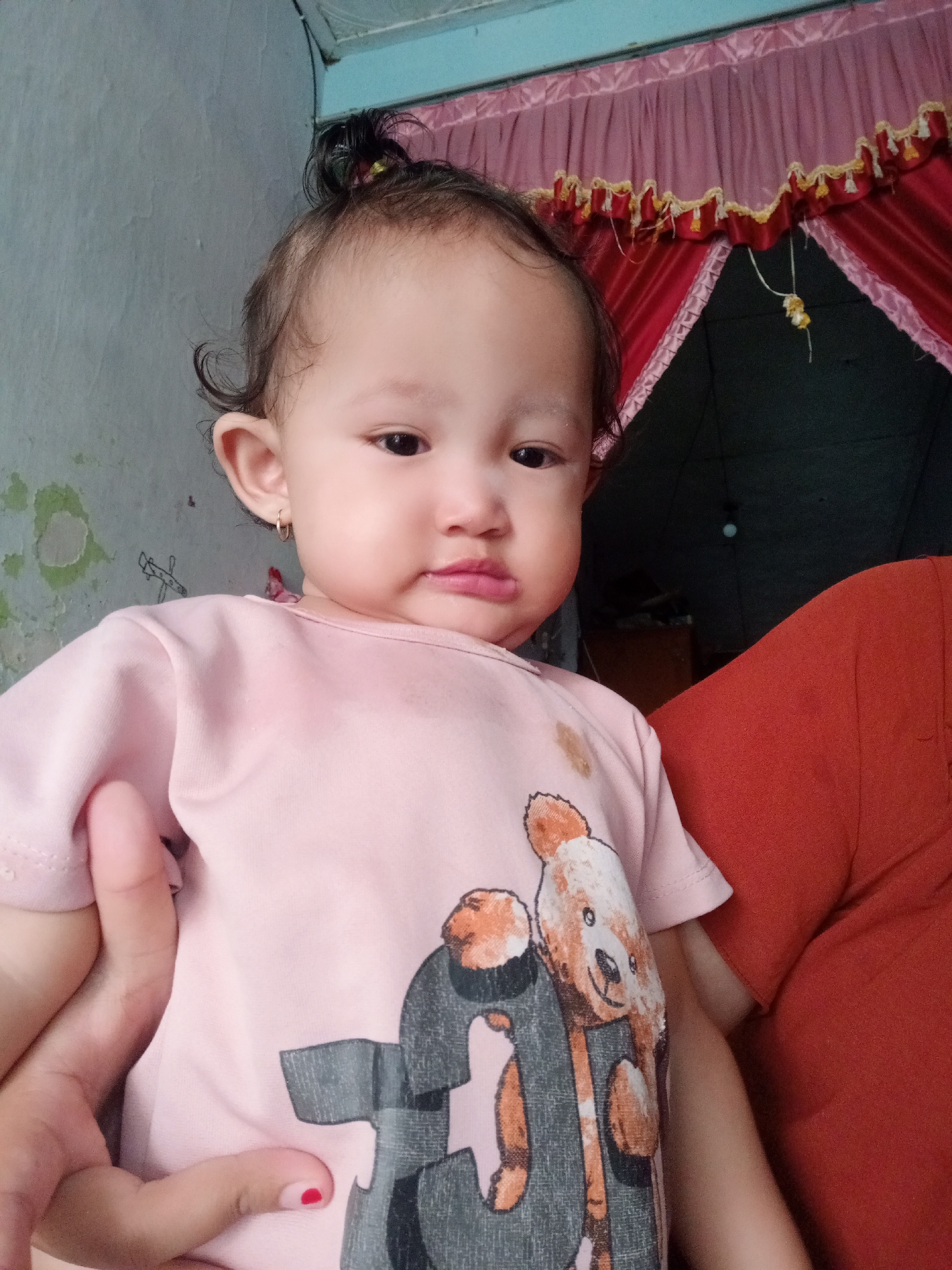 A baby is being held in someone's arms. The baby is wearing a pink shirt with a teddy bear graphic and small hoop earrings. The person holding the baby is wearing an orange shirt. The background shows a wall with some peeling paint, a partially visible blue ceiling, and a window with red and pink curtains with gold tassels. The location is Bungur, Indonesia.