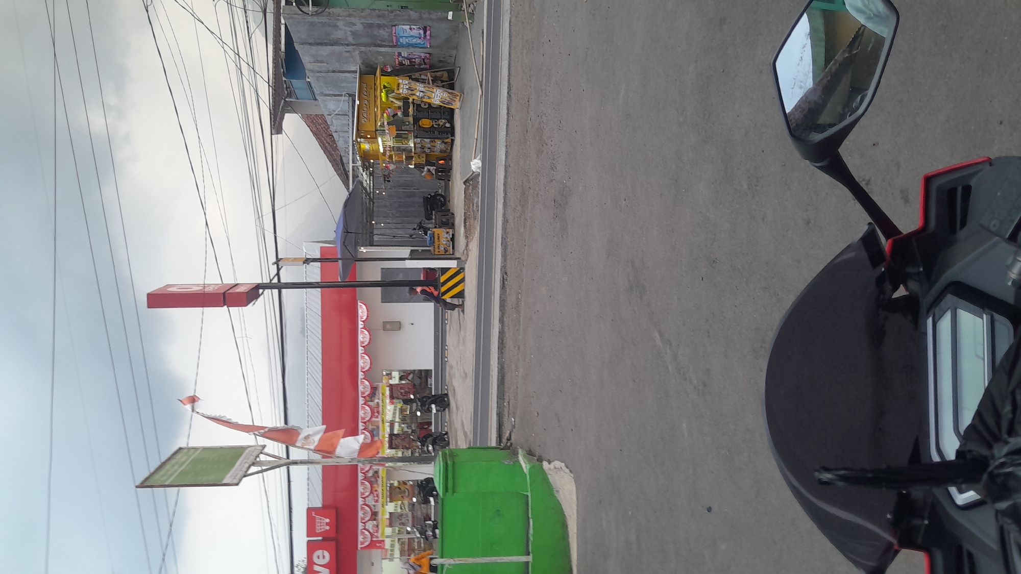 The image depicts a street scene in Subang, Indonesia. In the foreground, the handlebars and windshield of a red motorcycle are visible, indicating the photo was taken from the rider's perspective. The road surface is asphalt.
The middle ground shows a row of businesses. On the left is a brightly lit red building with a white facade and a prominent red sign with white lettering and a shopping cart icon. An Indonesian flag, partially unfurled and red and white, is attached to a pole in front of this building. A green barrier runs along the left edge of the frame, bordering the sidewalk. Several motorcycles are parked in front of the red building.
To the right of the red building, a smaller yellow food stall with a sign reading "Galaxy Local" is set up, featuring umbrellas, lighting, and various food items displayed. A person in an orange vest is seated on a low wall near the food stall. Another person is seen inside the food stall. A blue umbrella is open over a seating area next to the stall.
In the background, power lines crisscross the cloudy sky, and more buildings and structures are visible. The overall environment appears to be a commercial area in a town or city.