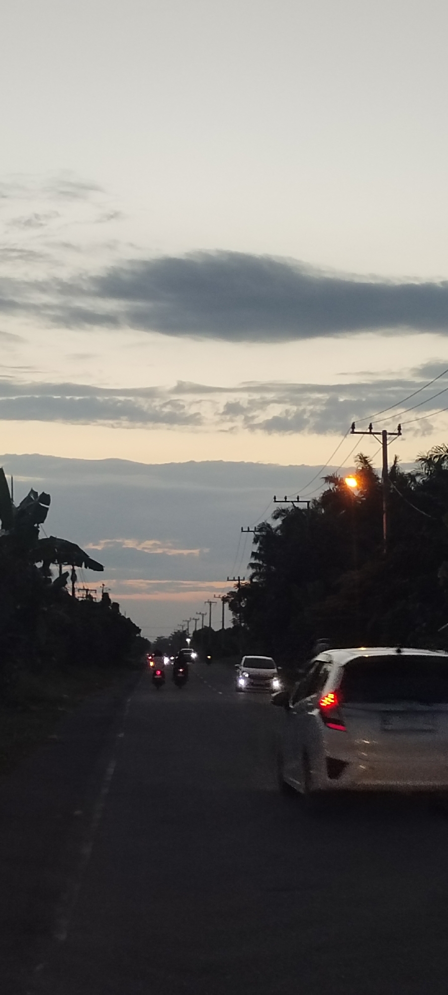 The image depicts a street scene in Kruengseupeng, Indonesia at dusk or dawn. The road extends into the distance, flanked by trees and utility poles. Multiple vehicles, including cars and motorbikes, are visible on the road. The sky is overcast with layered clouds. A street light is illuminated on the right side of the road.