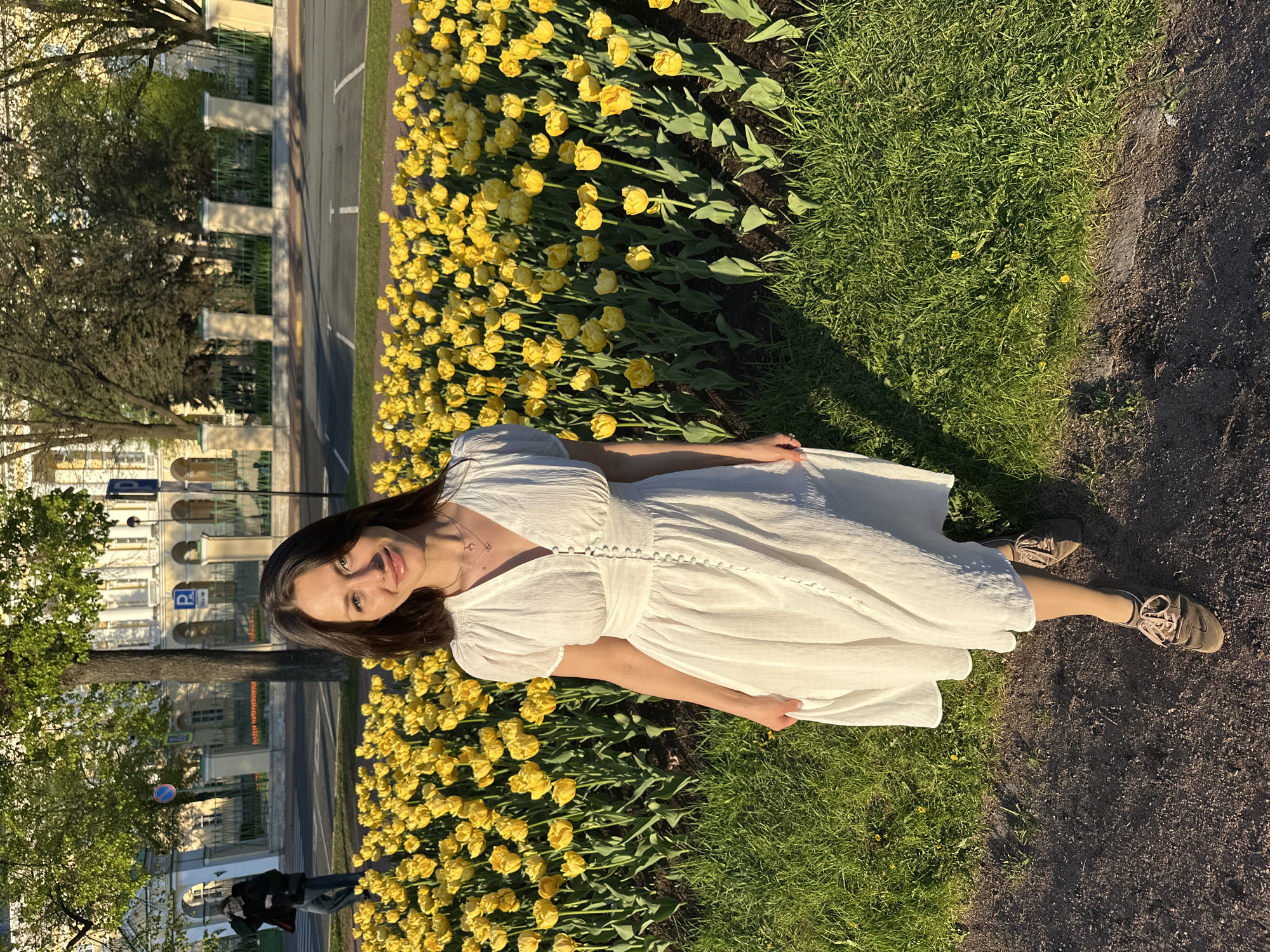A woman in a white dress stands in a field of yellow flowers, possibly tulips, in Saint Petersburg, Russia. A tree and buildings are visible in the background.