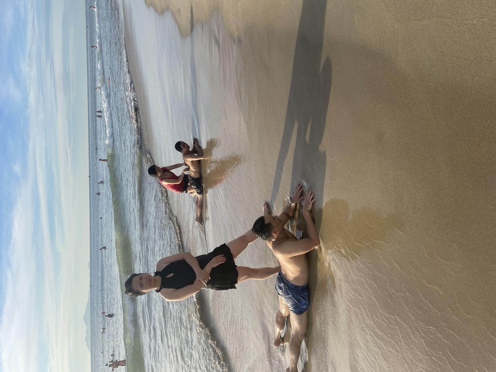 This is a photograph taken on a beach in Da Nang, Vietnam, likely during the late afternoon due to the lighting. In the foreground, a woman in a black dress stands on the wet sand, looking towards the camera with a neutral expression. Her arms are crossed. To her left, a man lies face down on the sand, his body partially submerged in shallow water. He is wearing blue patterned swim shorts. Further back, another man in a red tank top and dark shorts is positioned behind and slightly to the right of the woman, appearing to be interacting with someone on the ground. Behind him, a third man lies prone on the sand. The background shows the ocean with gentle waves and several people swimming or wading in the water. Distant hills or mountains are visible on the horizon. The sky is partly cloudy. The overall scene depicts a casual beach setting with people engaged in various activities.