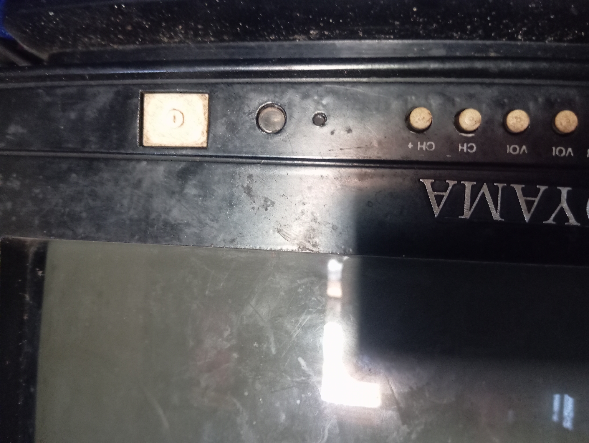 This is a close-up, eye-level, medium shot of the control panel of an old, analog television set. The television is predominantly black, with a matte finish that shows signs of wear and tear, including dust, scratches, and smudges. The screen is dark, reflecting bright lights from an unseen source, possibly windows or lamps, creating luminous rectangular shapes on the upper left and right portions of the screen.
The brand name "OYAMA" is prominently displayed in silver, capital letters across the bottom bezel of the television. Below the brand name, there is a row of controls. From left to right, these include:
* Four round, light brown buttons, arranged horizontally. The first two are labeled "VOL", and the next two are labeled "CH" and "CH+". The labels are in a subtle, darker font.
* A small, circular, dark button, possibly for power or a menu function.
* A larger, circular, metallic button, likely a volume or channel selector.
* A rectangular, beige-colored button with a circular symbol inside, possibly a power or menu button.
The area below the control panel is out of focus but appears to be a dark surface, possibly the stand of the television, with a visible accumulation of dust. The overall impression is that of a used, possibly vintage, electronic device. The scene is static and does not depict any people or actions.
The location is a residence in the city of Rejosari, Indonesia. This specific image focuses on the technological artifact, not the broader environment.