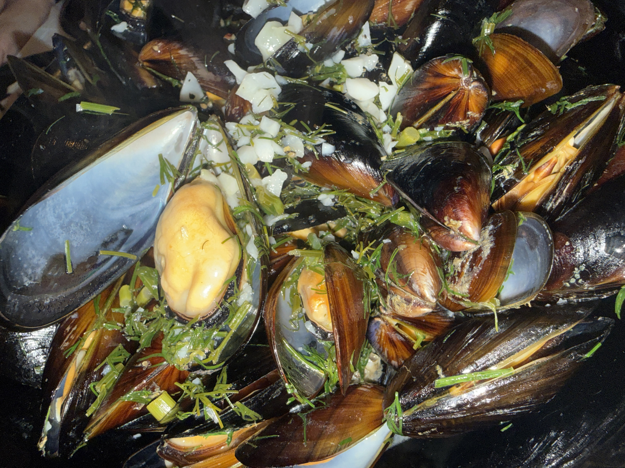A close-up, overhead shot captures a steaming pot of mussels, glistening with their juices and interspersed with chopped herbs, possibly dill and chives, and small pieces of white onion. The mussels are a mix of open and closed, their shells exhibiting a dark, iridescent brown hue. Some mussels reveal their plump, pale yellow flesh. The dish appears to be prepared for consumption, suggesting a culinary setting. The image does not contain any specific landmarks or indicators to pinpoint its location within Sozopol, Bulgaria.
