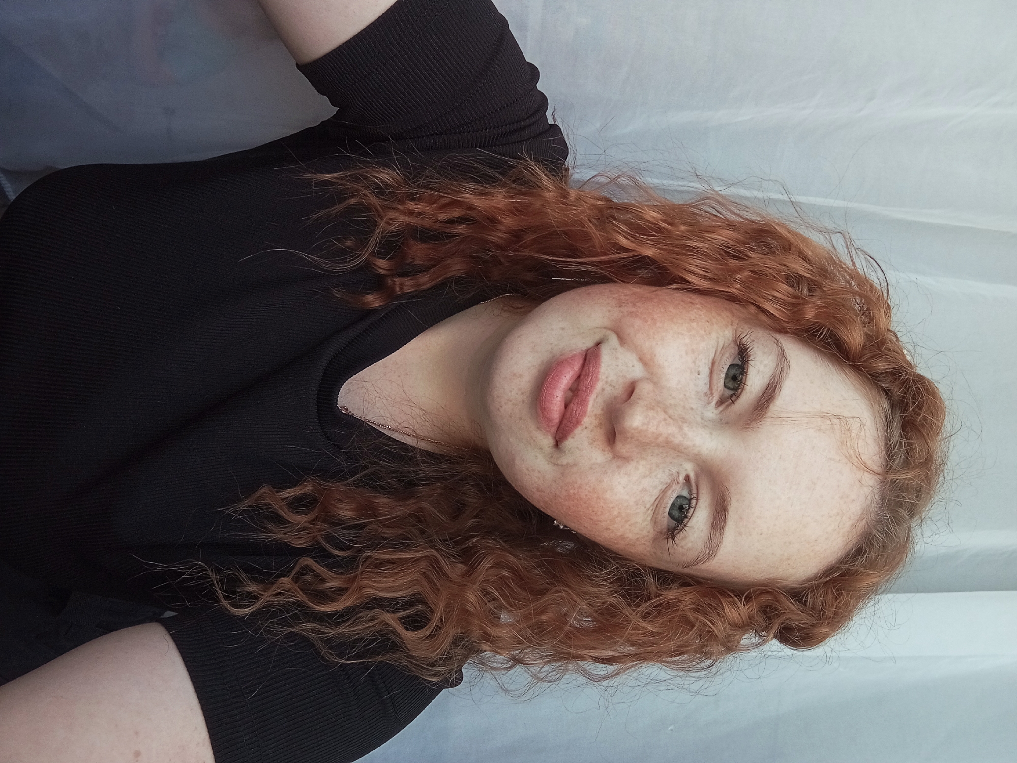A portrait of a fair-skinned young woman with red curly hair, freckles, and light-colored eyes is captured against a white, possibly draped, background. She is wearing a black top. No location in the city of Yurgamysh, Russia, is evident in the image.