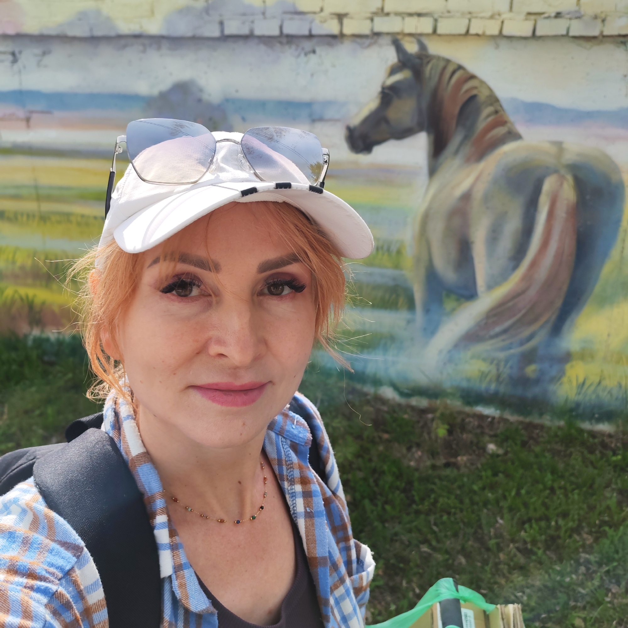 A fair-skinned woman with short, reddish-blonde hair wearing a white baseball cap with sunglasses perched on top is taking a selfie. She is wearing a blue and white plaid shirt over a dark-colored top, with a black backpack visible over her left shoulder. A delicate necklace with small, colorful beads is around her neck.
Behind her is a mural painted on a brick wall, depicting the rear view of a horse. The mural is set against a backdrop of green fields and a hazy mountain range. The ground at the base of the wall is covered in green grass.
The location is Chaikovsky, Russia.
