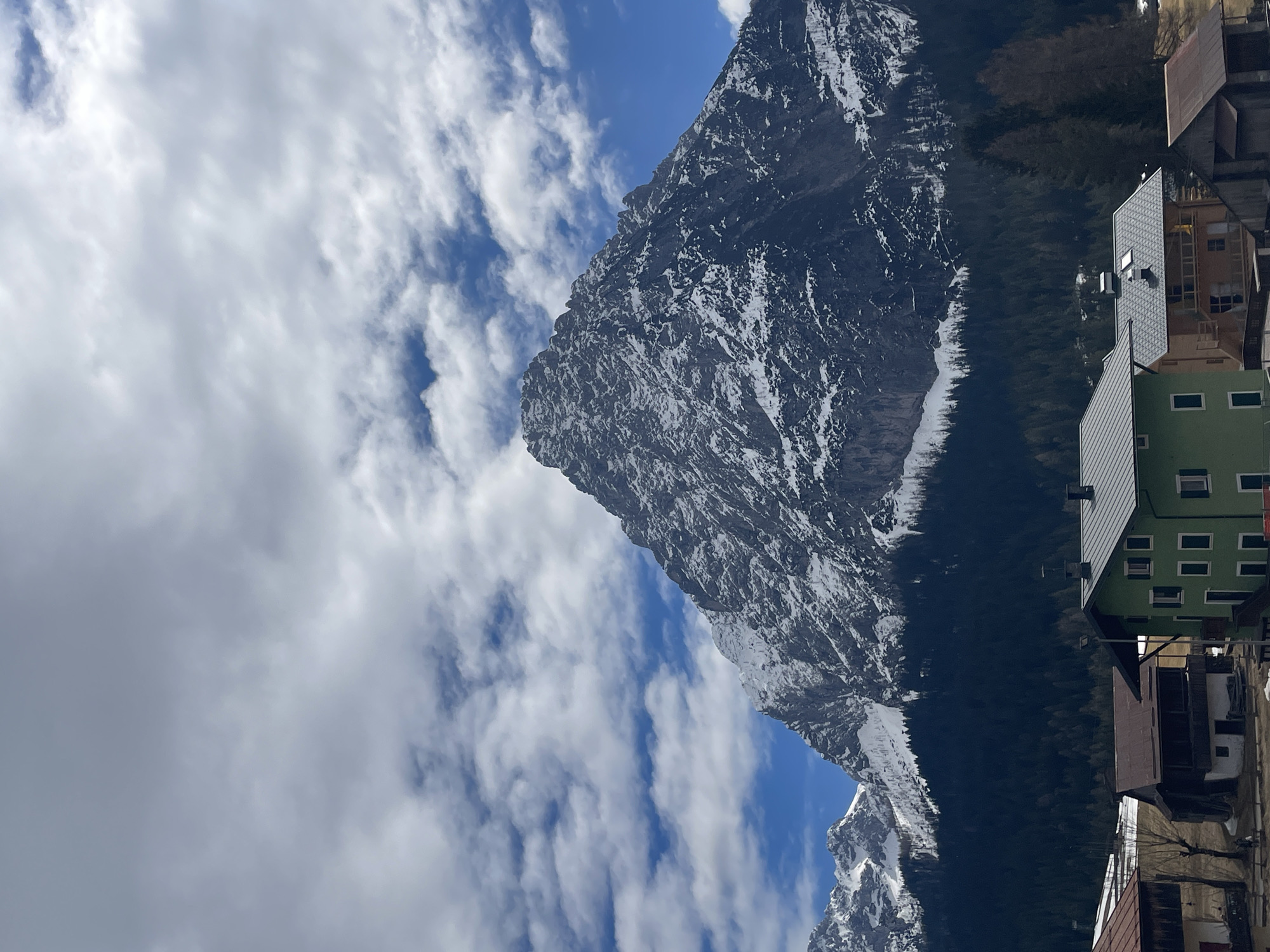 The image captures a view in the city of Sappada, Italy. In the foreground, there are buildings, one of which is green, suggesting an alpine village setting. The central focus is a large, snow-capped mountain, presumably part of the Dolomites, against a partly cloudy sky. The mountain's slopes are steep and textured, with visible rocky areas. The mountain is covered with a dense, evergreen forest that appears to ascend its lower slopes. The sky features scattered clouds and areas of clear blue, indicating a fair-weather day.