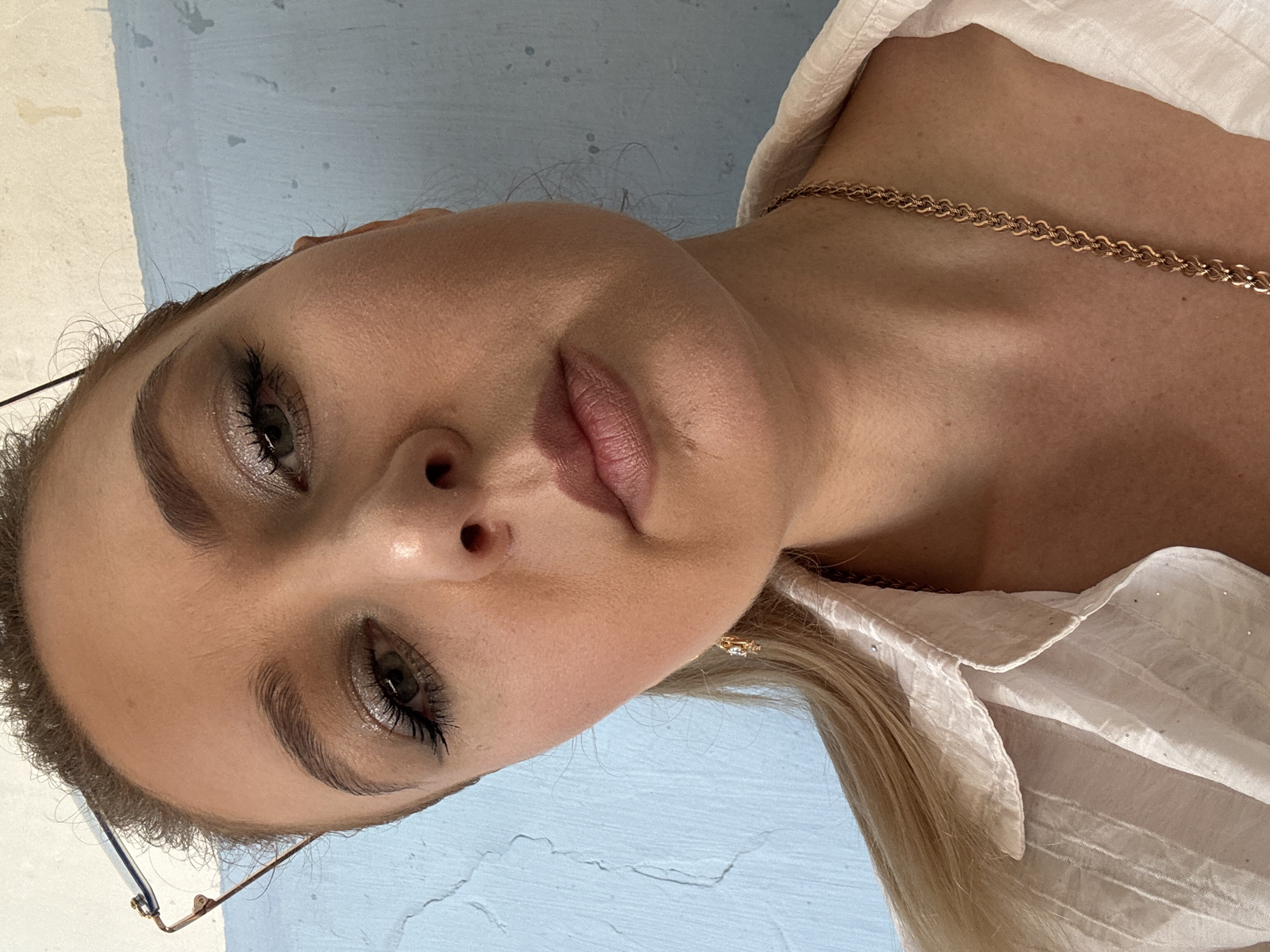 A close-up, waist-up, low-angle indoor portrait shows a young Caucasian woman with blonde hair pulled back, wearing a white button-down shirt and a gold chain necklace. She is looking towards the camera with a neutral expression. Her makeup includes glittery eyeshadow and mascara. She is wearing glasses perched on her head, and a small gold earring is visible. The background is a blue and white textured wall.