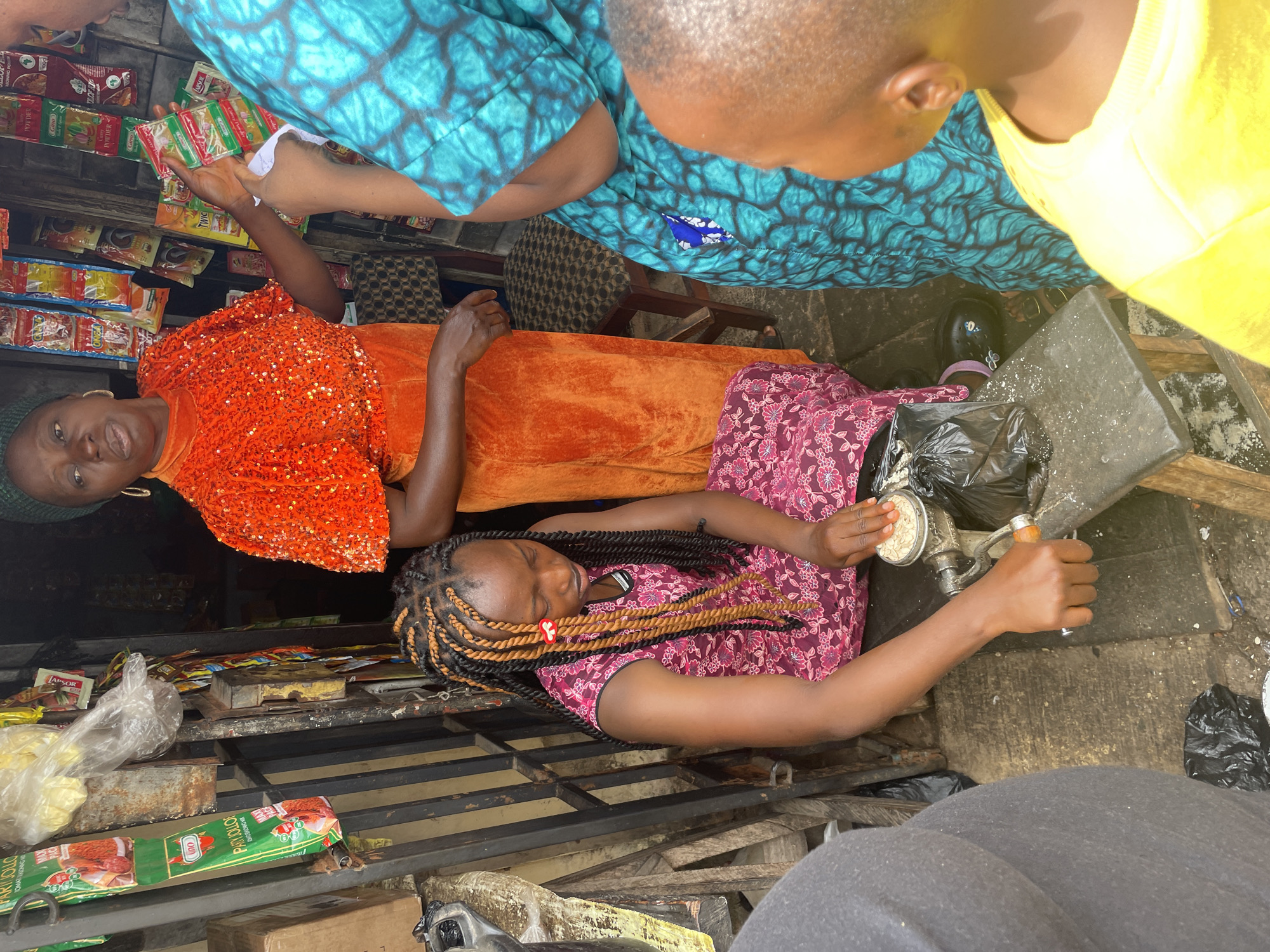 The image captures a vibrant scene in Jos, Nigeria. Three women are visible, two standing and one seated, seemingly engaged in a transaction near a stall or shop. The standing women are dressed in colorful, patterned clothing – one in a bright orange sequined top and a long orange skirt, and another in a turquoise blue outfit. They appear to be holding small packets or items from the stall. The seated woman is wearing a pink floral dress and has braided hair with orange accents. She is operating a grinding machine, processing some sort of grain or seed. The stall is stocked with various food items, including packaged seasonings and spices. The environment appears to be an outdoor market or commercial area.