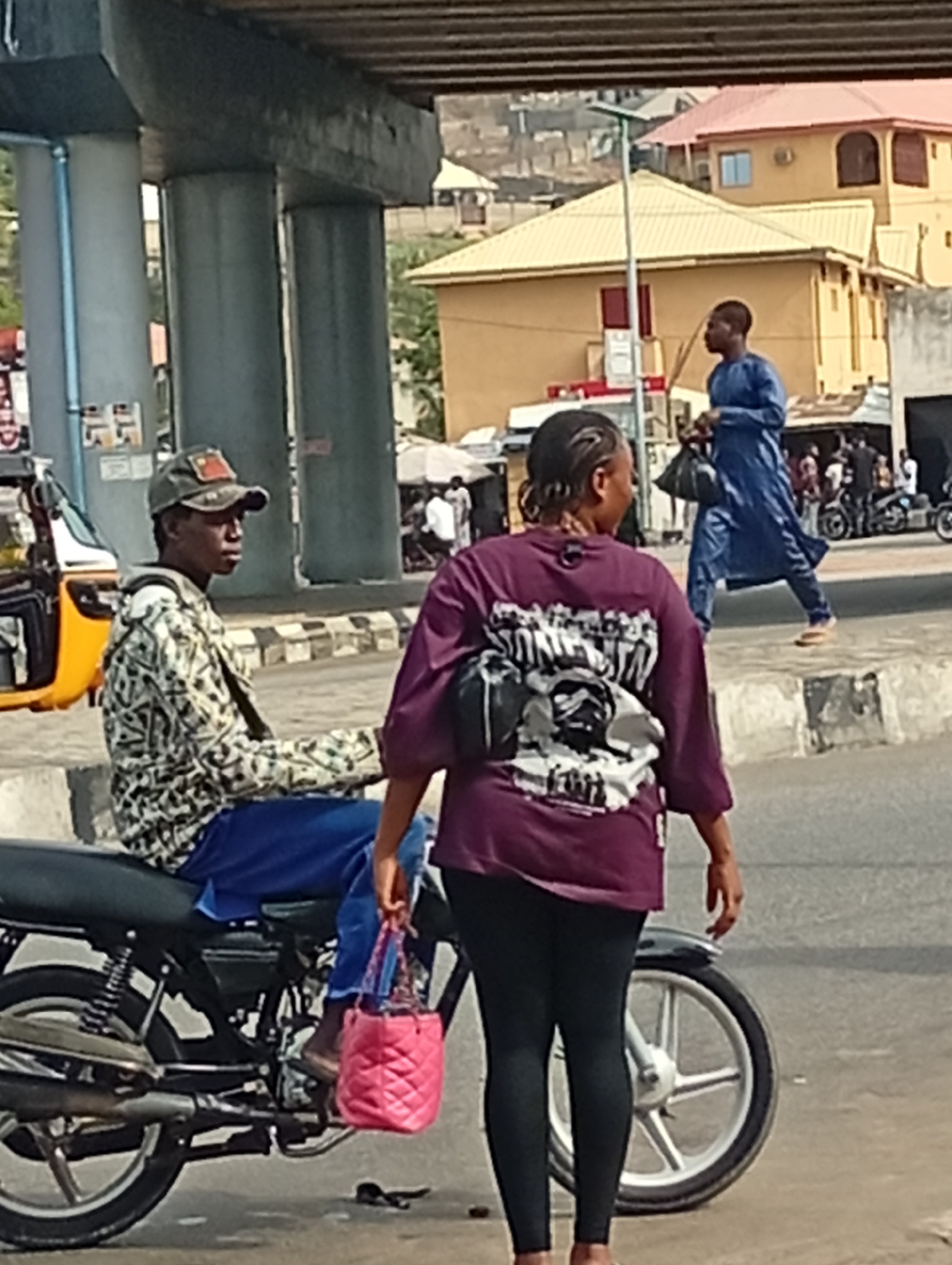 The image depicts a street scene in Lokoja, Nigeria. A man is sitting on a black motorcycle, wearing a cap, patterned jacket, and blue pants. He is looking to the left. A woman in a maroon t-shirt and black leggings stands to the right of the motorcycle, holding a pink purse. A man in a blue robe walks across a raised crosswalk in the background. The background includes buildings, other pedestrians, and a yellow tricycle. The scene appears to be near a concrete overpass.