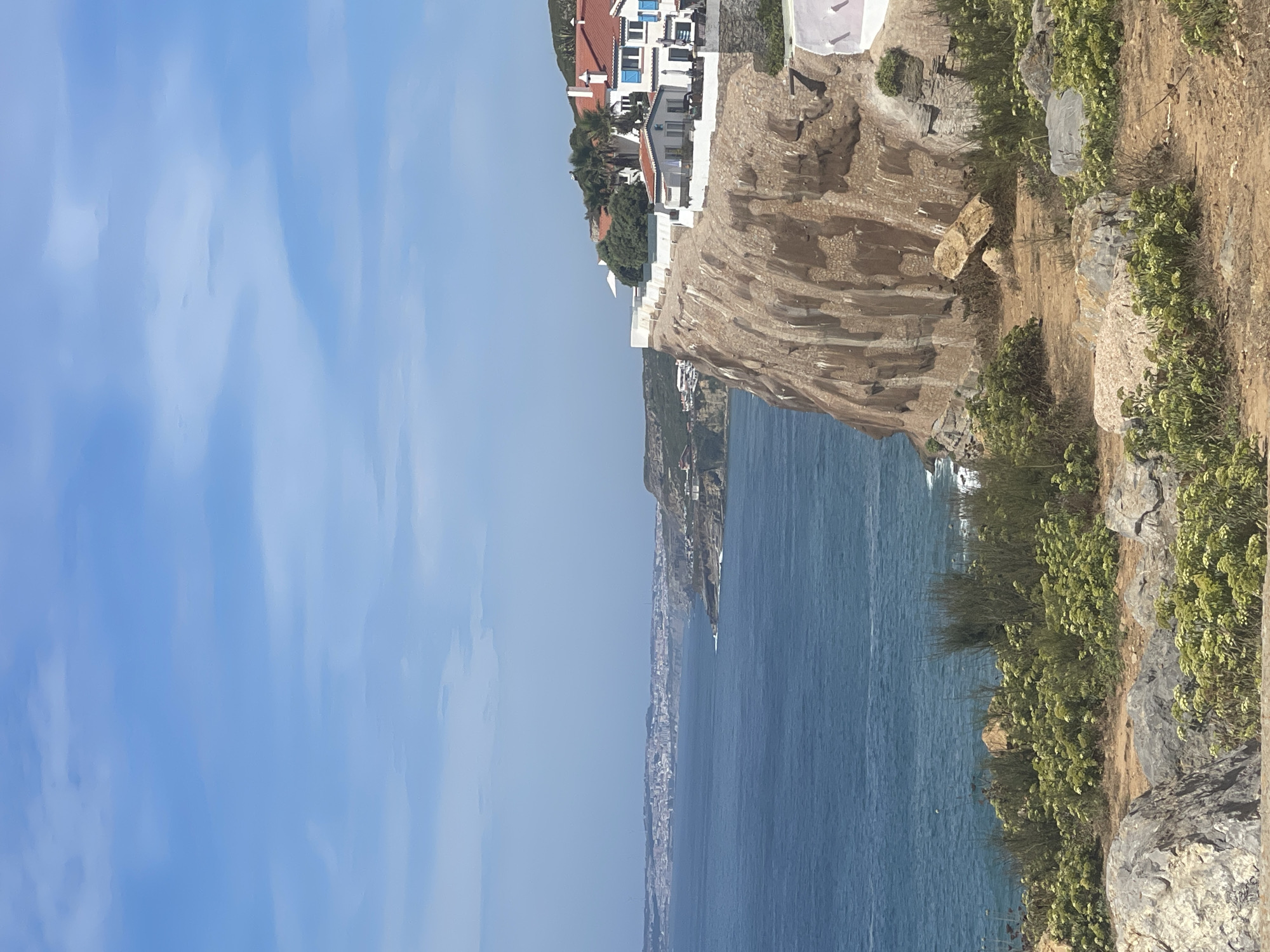 A scenic view of the coastline in Colares, Portugal, shows a rugged cliff face overlooking the Atlantic Ocean. On top of the cliff, a row of white houses with red-tiled roofs and blue shutters is visible. The houses are situated along a white-walled promenade. The ocean is a deep blue, with waves gently breaking against the shore. The foreground is characterized by rocky terrain and sparse vegetation, including green bushes and wildflowers. In the distance, a sprawling city can be seen along the coastline. The sky is a clear blue with scattered white clouds.