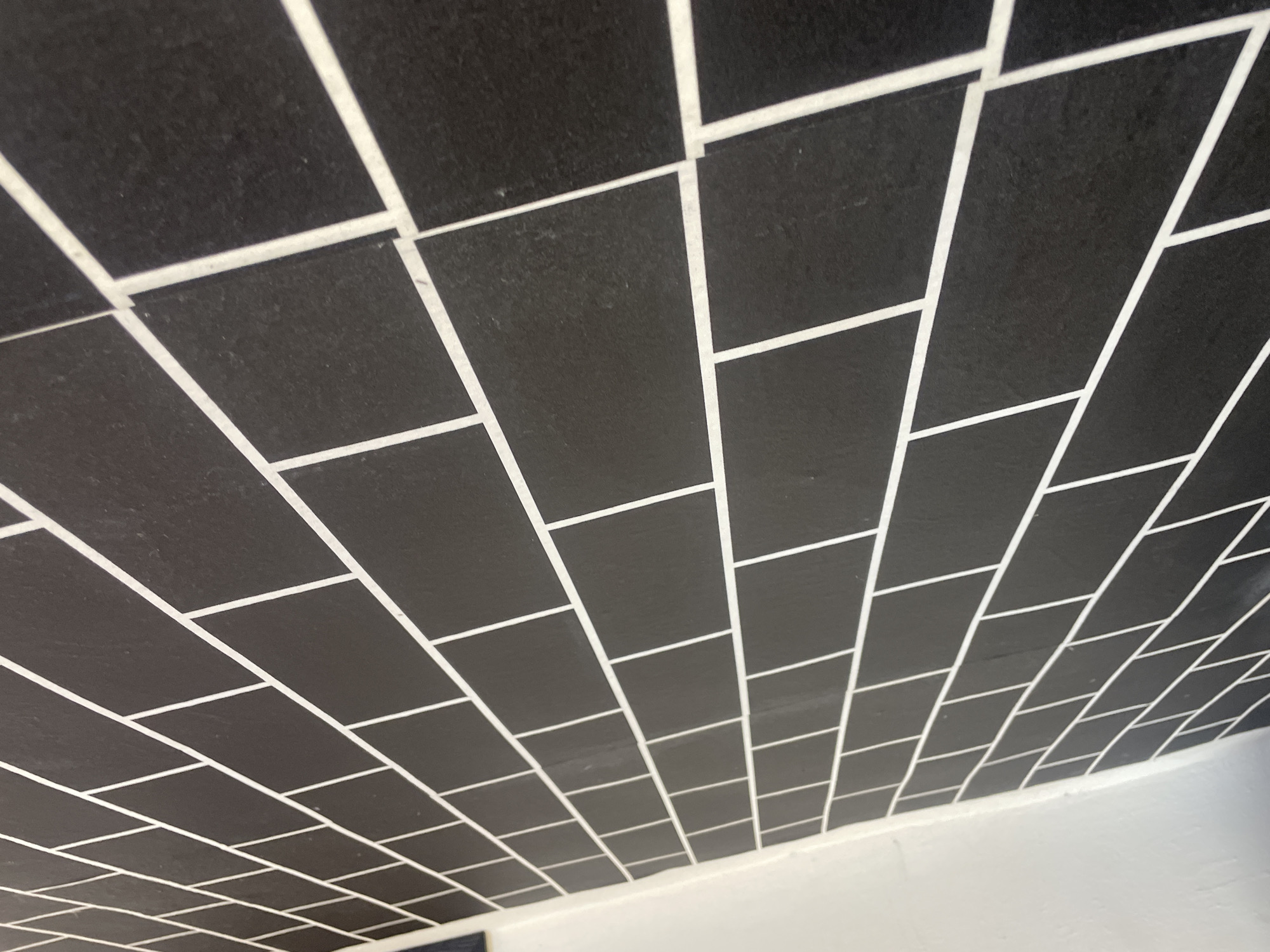 The image shows a close-up of a dark gray tiled wall with thin white grout lines. The tiles are rectangular and arranged in a staggered pattern, resembling a brick wall. The perspective is from a low angle, looking up and to the right, with the wall extending out of the frame. The wall appears to be an interior surface, possibly in a home or commercial building. The lighting is even, and there are no shadows to indicate a specific light source. The image does not provide any information about the location in the city of Jalingo, Nigeria, or any people, subjects, or actions. It is a static depiction of a wall's surface.
