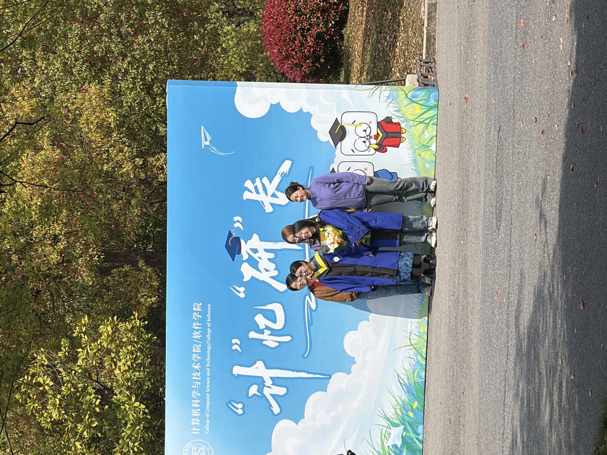 The image shows a group of four young women posing in front of a large sign. The sign is light blue and has the words "计算机科学与技术学院/软件学院 College of Computer Science and Technology/College of Software" along the left side, as well as large white stylized Chinese characters. A cartoon graduate is depicted on the upper right. The women are dressed casually, with some wearing graduation robes. They are standing on a paved surface, likely a street or sidewalk. Trees are visible in the background, suggesting an outdoor location. A dark red shrub is also visible. The image appears to be taken in the city of Jiangning District, China.