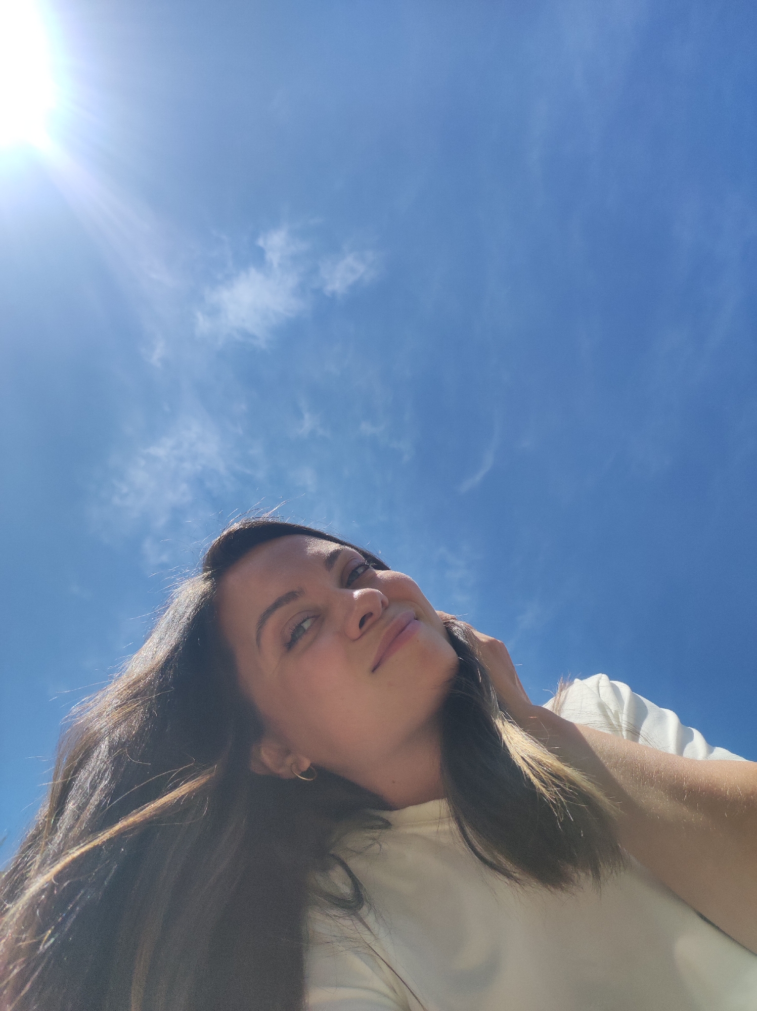 The image is a low-angle selfie of a woman against a bright blue sky with scattered clouds and a visible sun flare. She is fair-skinned with long, dark brown hair and is wearing a light-colored shirt. Her head is tilted, and she is looking slightly to the right of the camera. Her left arm is raised, with her hand possibly resting on her neck or head. The specific location within the city of Chaikovsky, Russia, cannot be determined from the image alone.