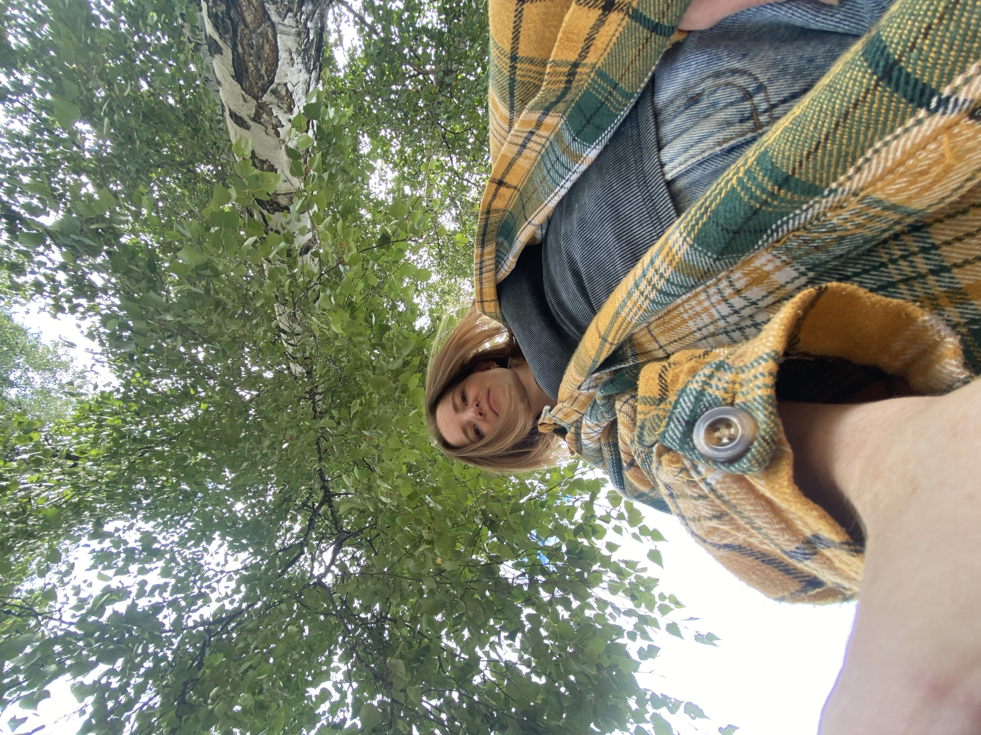 An upward-facing shot shows a woman wearing a yellow and green plaid shirt over a dark gray top and blue jeans. She is looking up at the camera, her blonde hair slightly windswept, framed by the dense green leaves of a birch tree overhead. The rough texture of the birch bark is visible on the right side of the frame. The background is a bright, overcast sky peeking through the foliage, suggesting an outdoor setting in Chaikovsky, Russia, likely a park or natural area given the prominent birch tree.