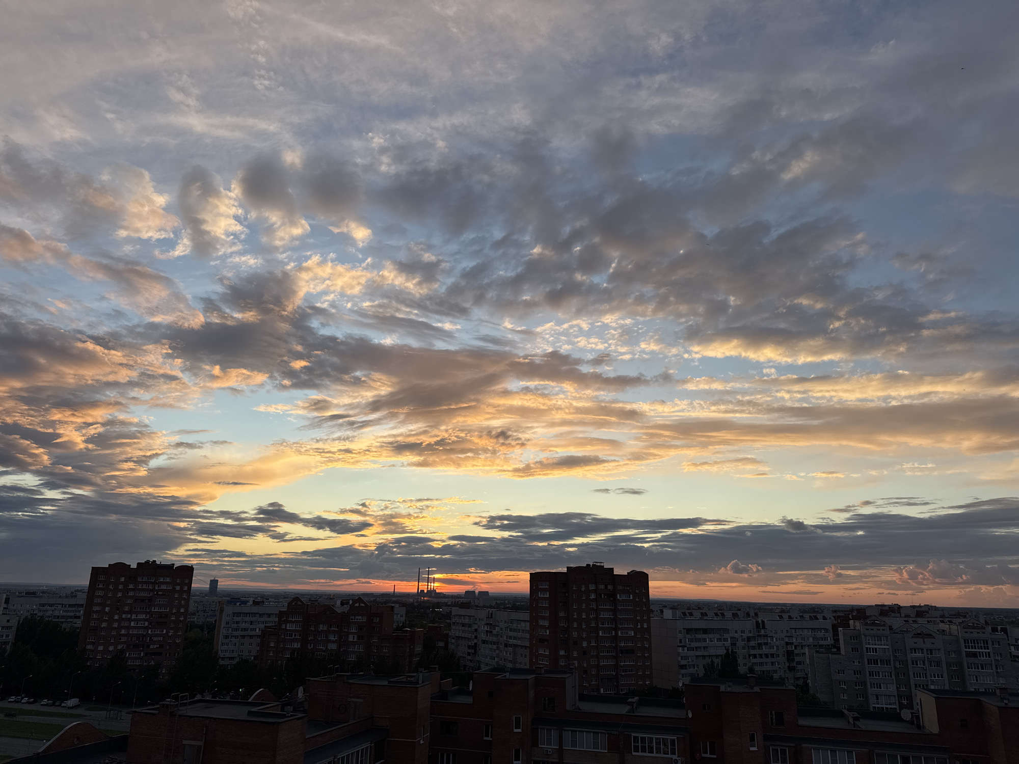The image depicts a city skyline at sunset. The sky is filled with dramatic clouds, illuminated by the warm hues of orange, pink, and gold, contrasting with areas of blue and grey. Silhouetted against the colorful sky are numerous apartment buildings of varying heights and architectural styles, creating a dense urban landscape. In the distance, the silhouette of a tall, slender building is visible, along with what appear to be industrial structures or power lines. Trees line some of the streets, and the rooftops of lower buildings are also visible in the foreground. The overall impression is one of a vast, populated urban area experiencing the beauty of twilight.