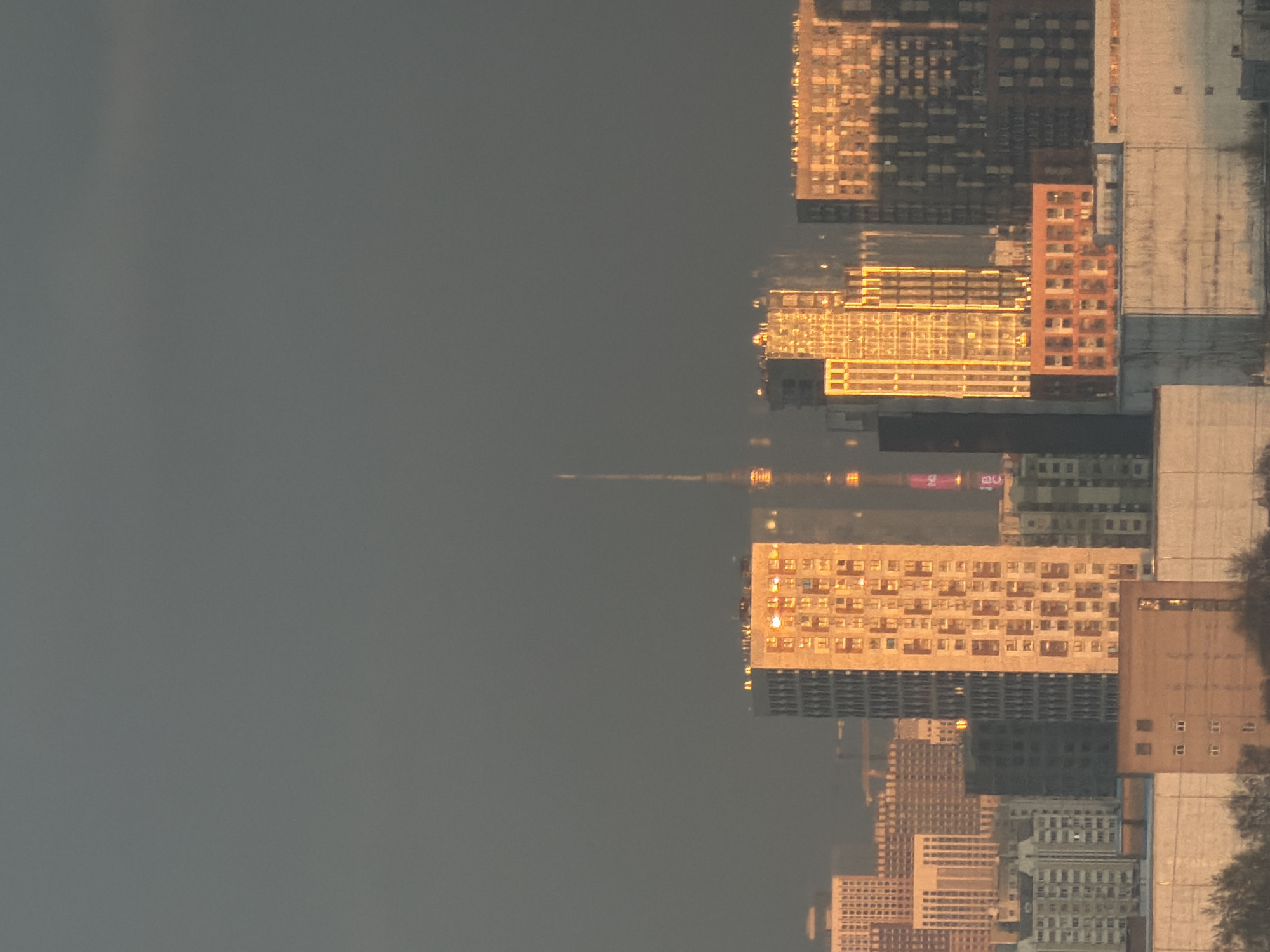 The image shows a reflection of a cityscape in a body of water, likely a lake or river. The buildings are various heights and colors, with some appearing to be residential and others possibly commercial. There are no people visible in the frame. The environment seems to be dusk or dawn, judging by the lighting. The image is described as being a reflection in the city of Khimki, Russia.