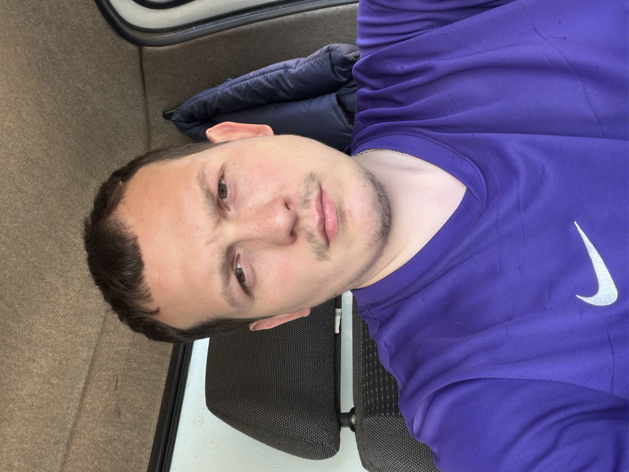 A young man with light skin, dark brown hair, and stubble on his chin and jawline, along with a faint mustache, is depicted in a rotated selfie. He has brown or hazel eyes and several freckles visible on his nose and cheeks. He wears a bright purple short-sleeved shirt featuring a white Nike swoosh logo on the right chest area. A thin silver-colored chain is visible around his neck.
The man appears to be seated or reclining, with his head resting near a dark, textured headrest or seat back. A white tag with the number "03" is faintly visible on the side of this seat element. Behind his head and shoulder, a dark blue padded jacket or similar item is present. The background to his left and above consists of a textured, light brown or beige surface, possibly carpeting or an upholstered interior wall of a vehicle or a small room. The overall lighting suggests it is daytime.
The image captures a direct, neutral gaze from the subject, who is likely taking the photograph himself. The setting is indoors, and given the context, it is located in Chaikovsky, Russia.
