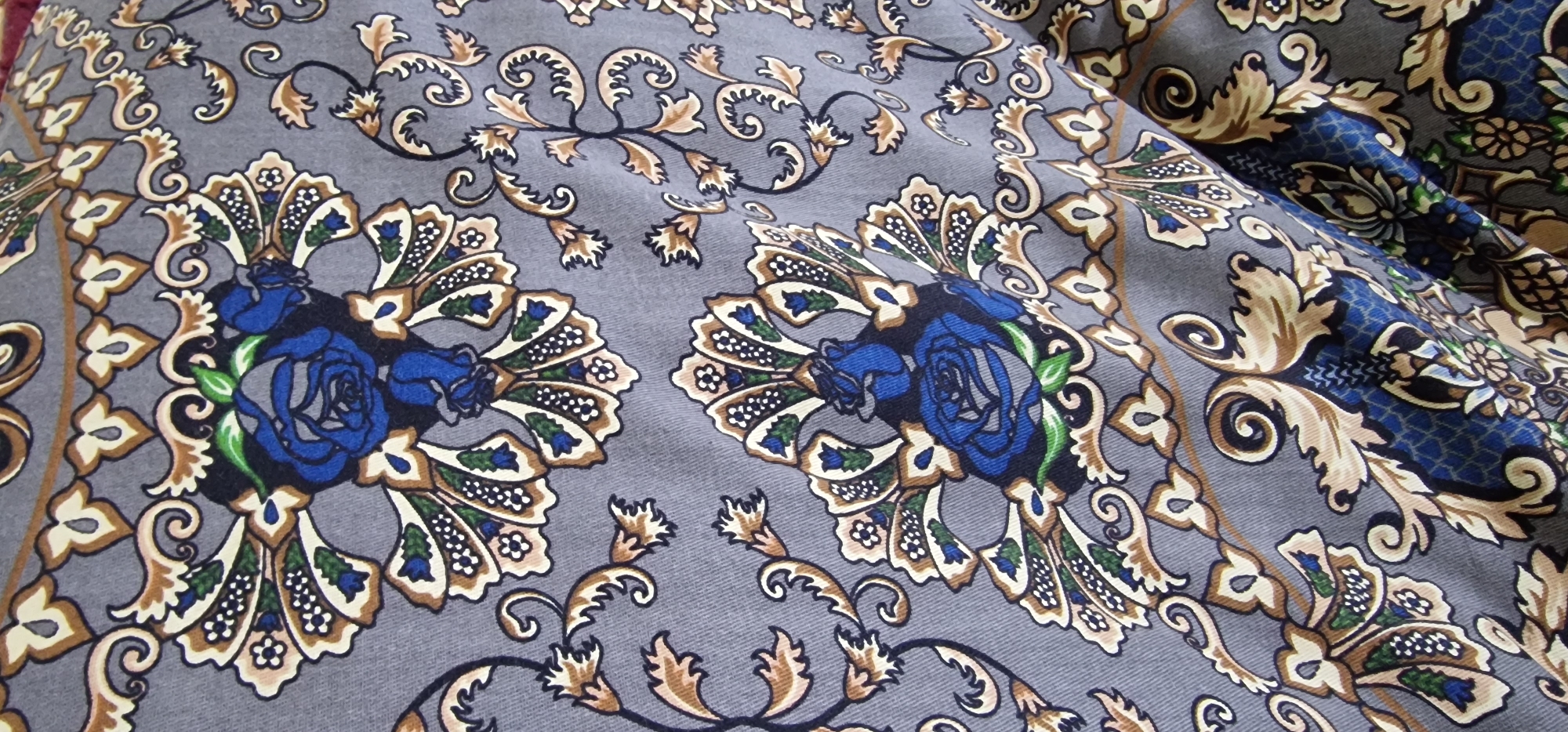The image displays a close-up of a grey fabric with an intricate, repeating floral and paisley pattern. The pattern features stylized blue roses, along with elements in shades of brown, cream, and green. The fabric appears to be folded or draped, creating shadows and highlights that emphasize its texture. There are no people, actions, or interactions visible in the image. The scene is a close-up of textile material. This image does not contain any elements that would allow for the determination of its location in Karshi City, Uzbekistan.