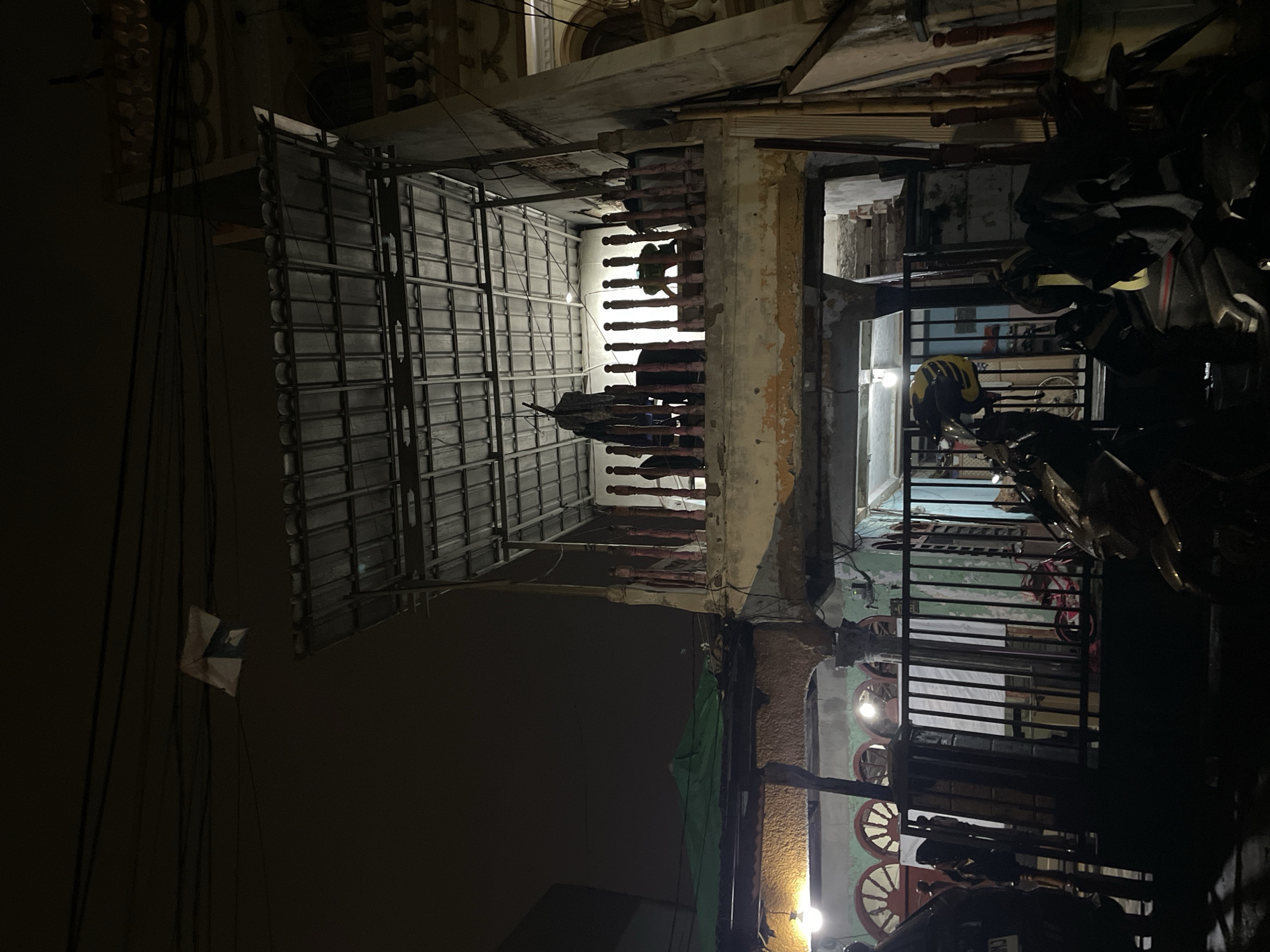 The image shows a building at night in North Jakarta, Indonesia. The building's exterior is weathered, with exposed elements and deteriorated paint. An open-air section under a corrugated metal roof houses clothing or textiles hanging to dry. A partial view into the building reveals objects, including bicycles and other items. Parked motorcycles obscure another area of the building. Power lines with a small flag or marker are visible against the night sky.