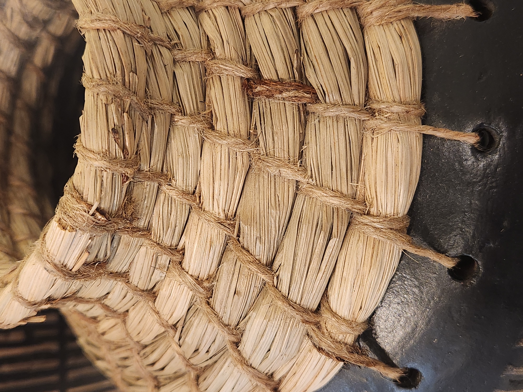 The image is a close-up, angled shot of the woven straw upper portion of a black ceramic vase. The straw is tightly bound with natural twine in a crisscross pattern. The twine also hangs in loose strands from the bottom edge of the straw, passing through small, dark holes in the black ceramic base of the vase. The straw appears dry and textured, with some pieces slightly frayed. The ceramic base has a matte, dark finish and is partially visible at the bottom and left side of the frame. The background is dark and out of focus, suggesting an indoor setting. The image does not depict any people or specific locations within Maitland, United States of America.