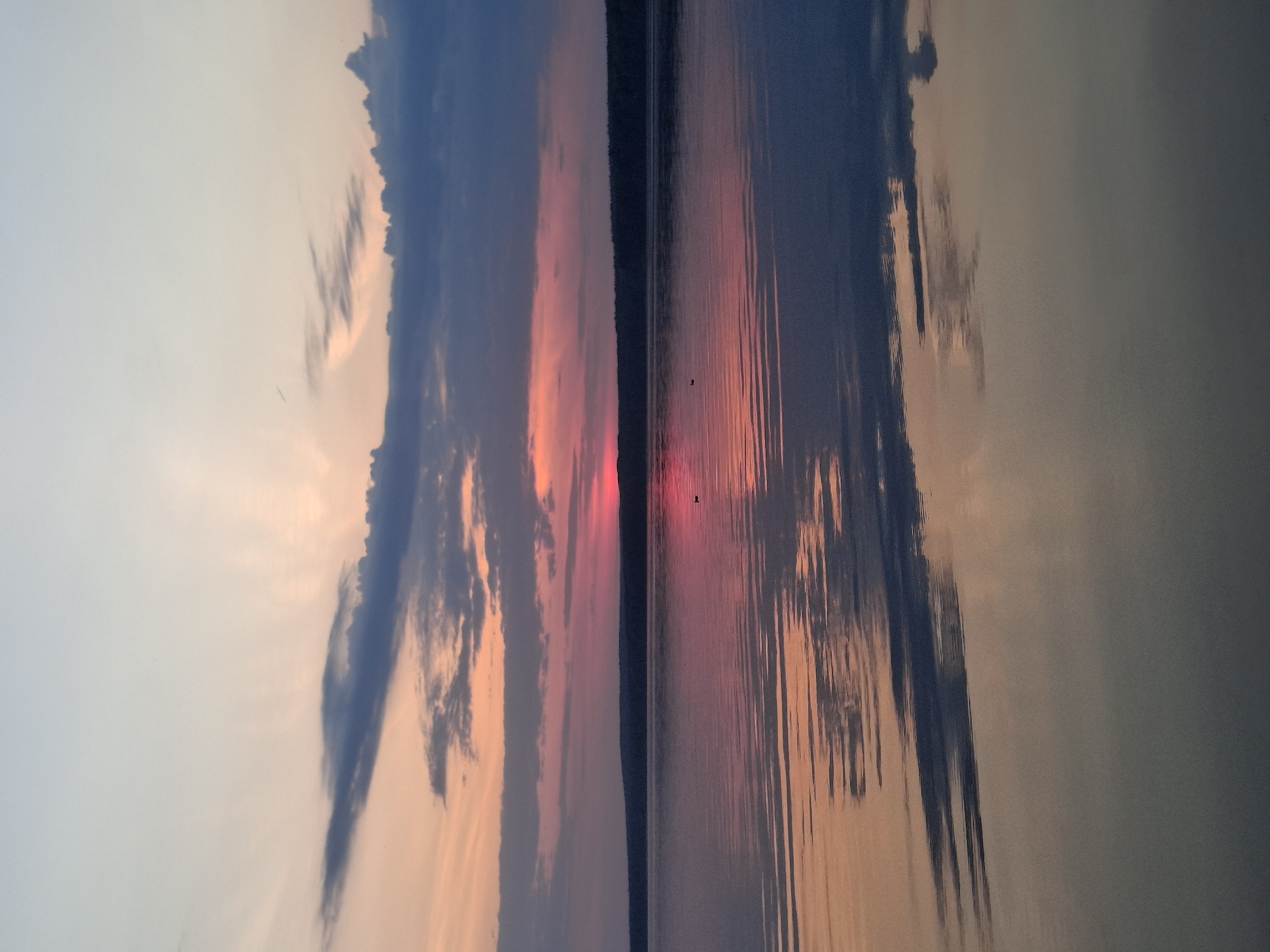The image shows a sunset over water, likely a lake or river. The sky is a mix of orange, pink, and blue hues, with dark clouds on the left and right sides. These colors are reflected in the water, creating a mirror-like effect. The horizon line has a thin, dark strip of land, possibly trees. The water is calm with subtle ripples. In the center of the water, near the horizon, there is at least one small dark spot that might be a bird or object floating in the water. The image is rotated, so the sky is on the bottom. It depicts a tranquil environment. While the location is given as Chaikovsky, Russia, specific identifiable landmarks are not present in the image.